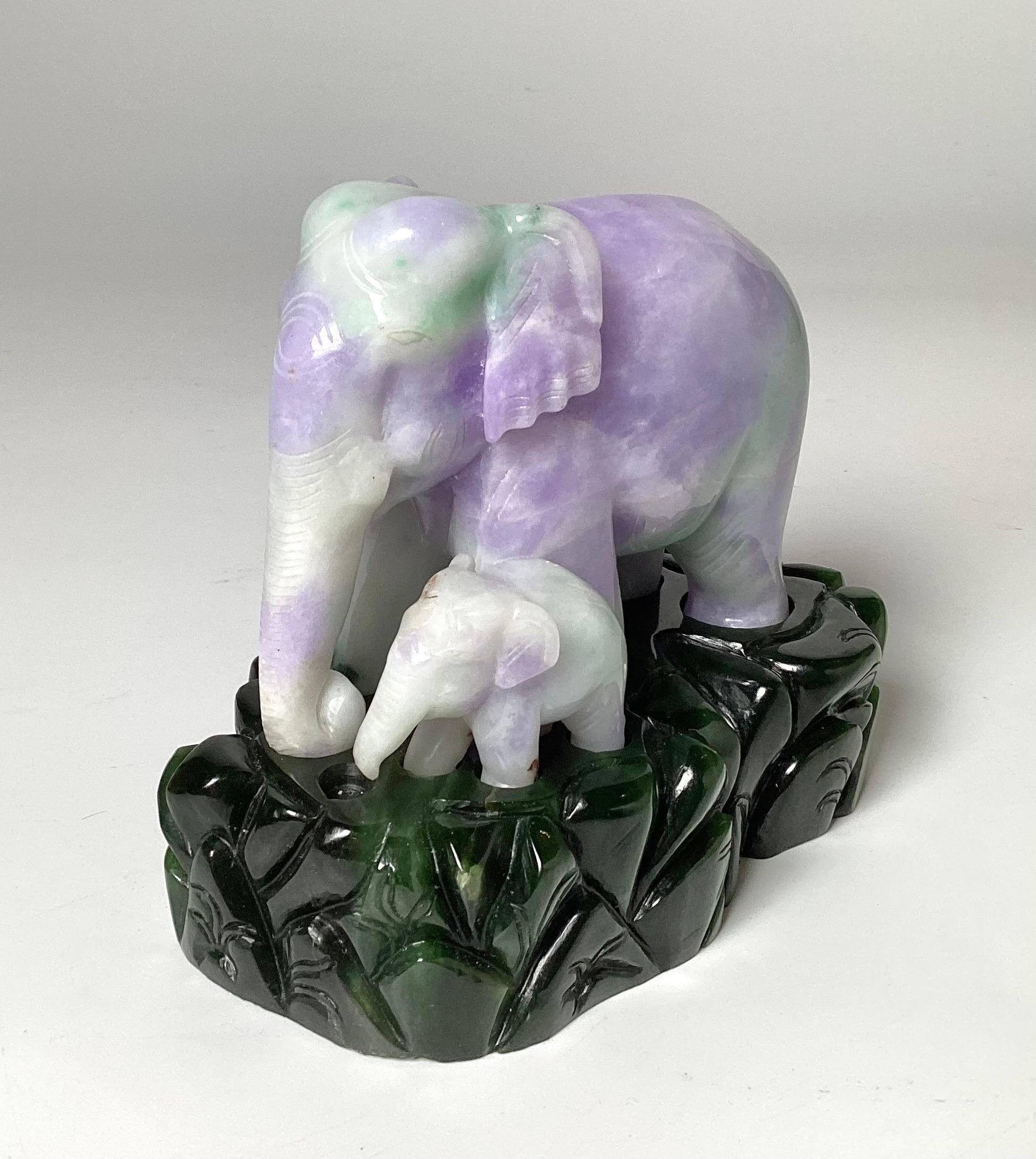 Charmant éléphant en jadéite sculpté sur une base en jade épinard. L'éléphant sculpté à la main en jade vert pomme et lavande représente une mère et son petit. Le socle vert foncé est séparé et la figurine repose dessus. 5 pouces de large, 4 pouces