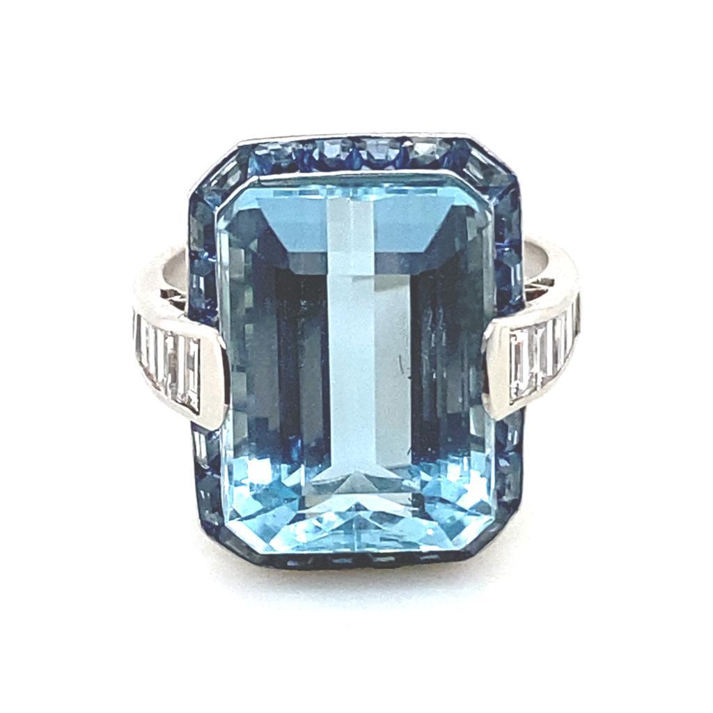 A vintage aquamarine, sapphire and diamond platinum cocktail ring, circa 1960.

Set to its centre with an emerald cut aquamarine of 10.13cts approximately in a handmade fine platinum bezel within a calibré  sapphire cluster frame, with bezel set