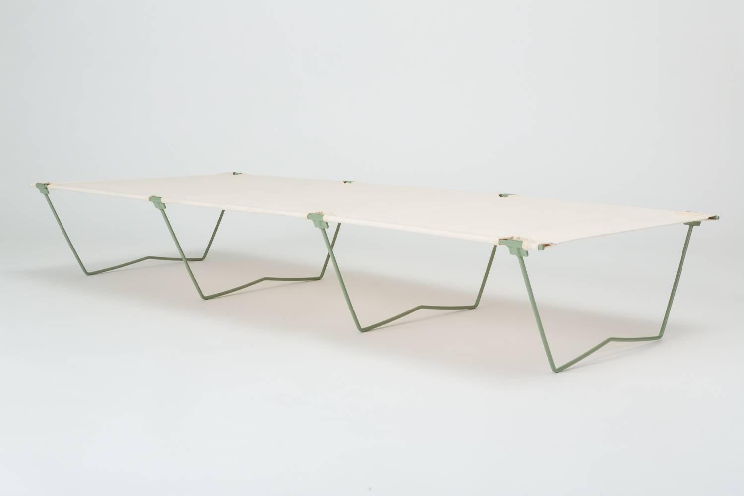 Similar to the collapsible army cot used by the British Army during World War II, this modernist cot has a steel frame powder-coated in a glossy sage green. Four bent legs support the bed from beneath and “clip on” to the frame. A stretched sling in