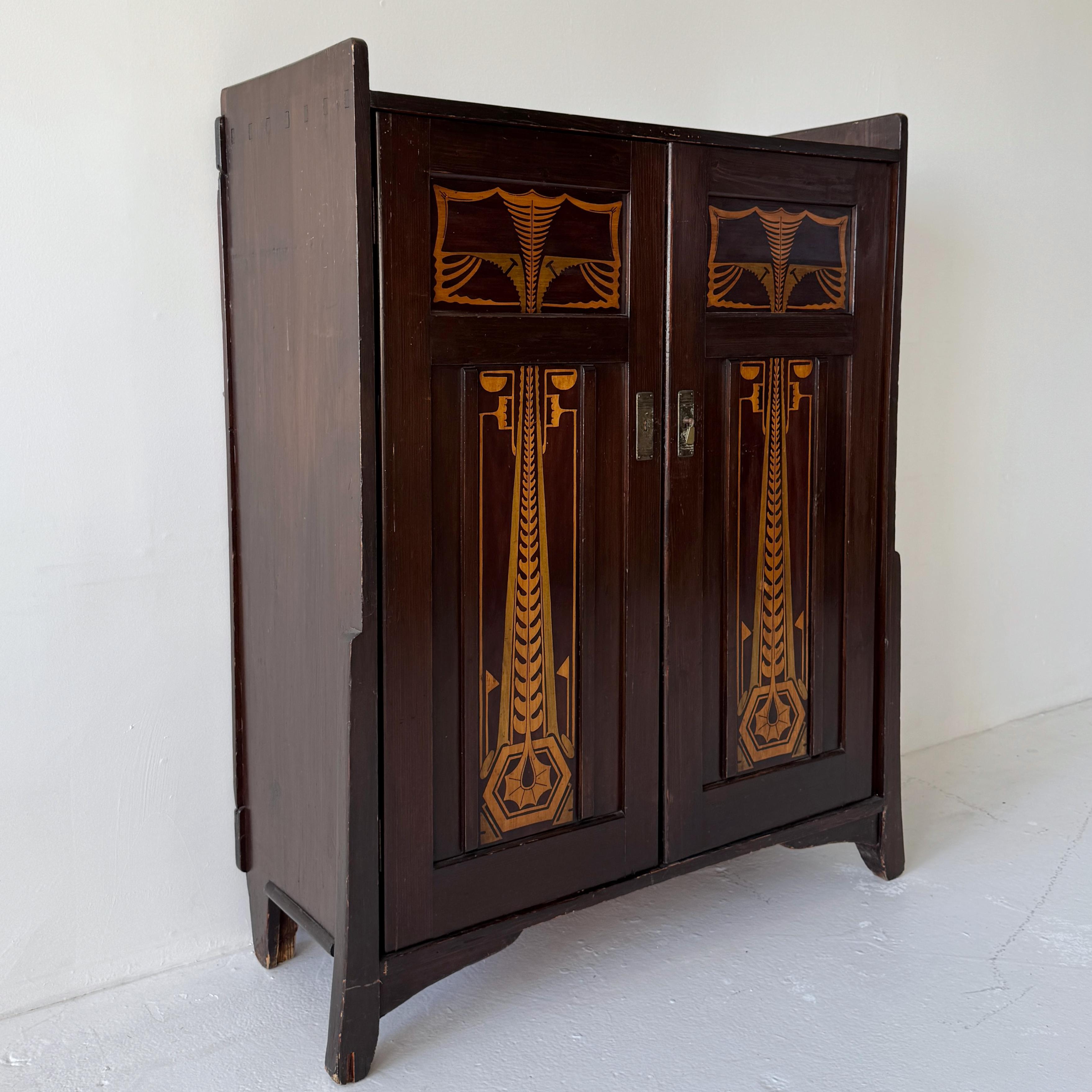 Early 20th Century Art Deco cabinet featuring striking Art Nouveau–inspired stained wood motifs on the double doors. The geometric and organic detailing showcases fine craftsmanship of the era. The cabinet opens to reveal four interior shelves,
