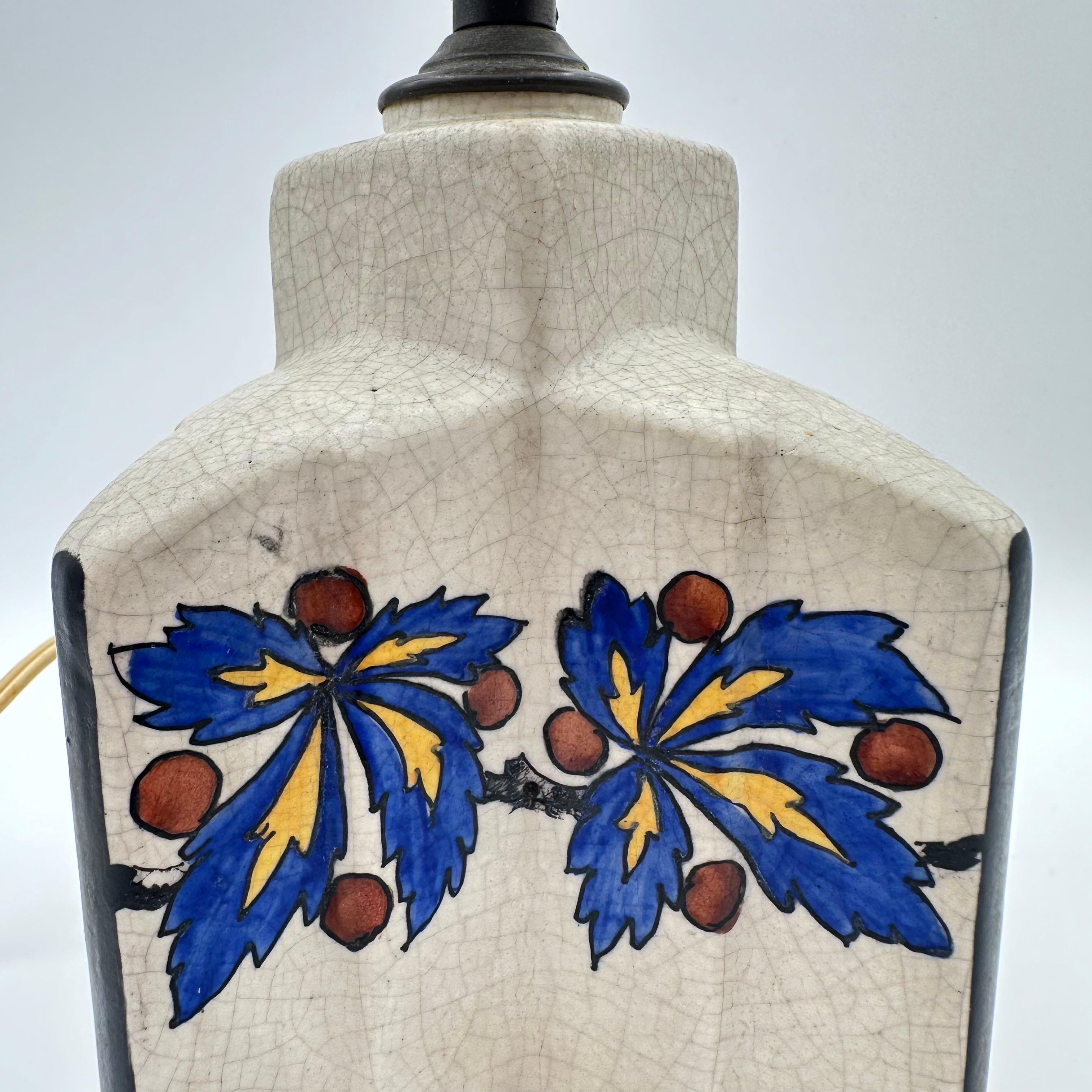 Vintage Art Deco table lamp by Antoine Dubois. Hand glazed ceramic with a motif of blue, yellow and deep red holly leaves and berries set atop an off white or egg shell colored craquelure glaze. A stepped four pointed star shape, with black lines