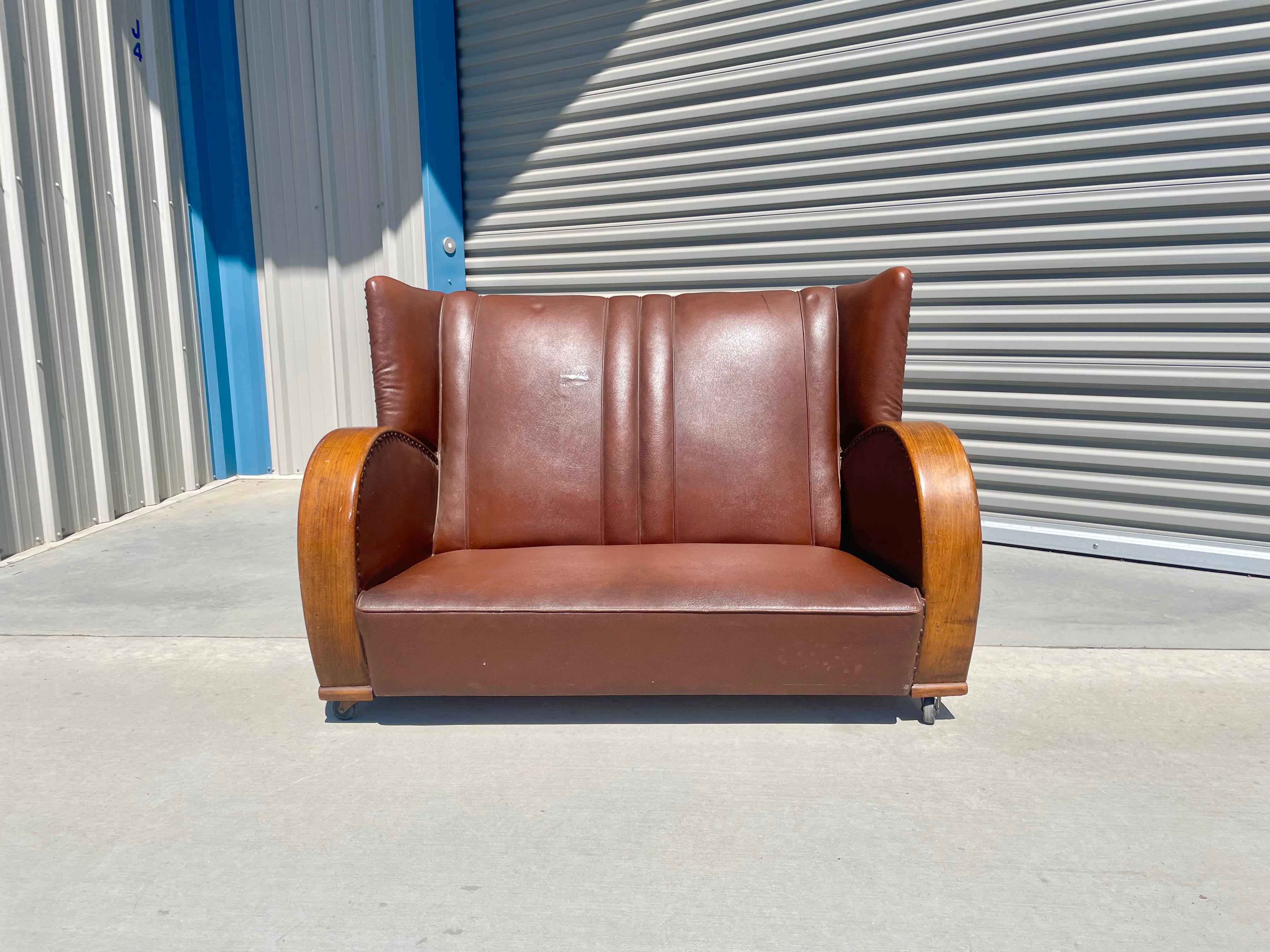 Vintage art deco leather loveseat manufactured in the united states, circa 1970s. This beautiful loveseat features two barrel form armrests that lead down, giving it a tremendous distinctive design for its era. The loveseat also features wheels on