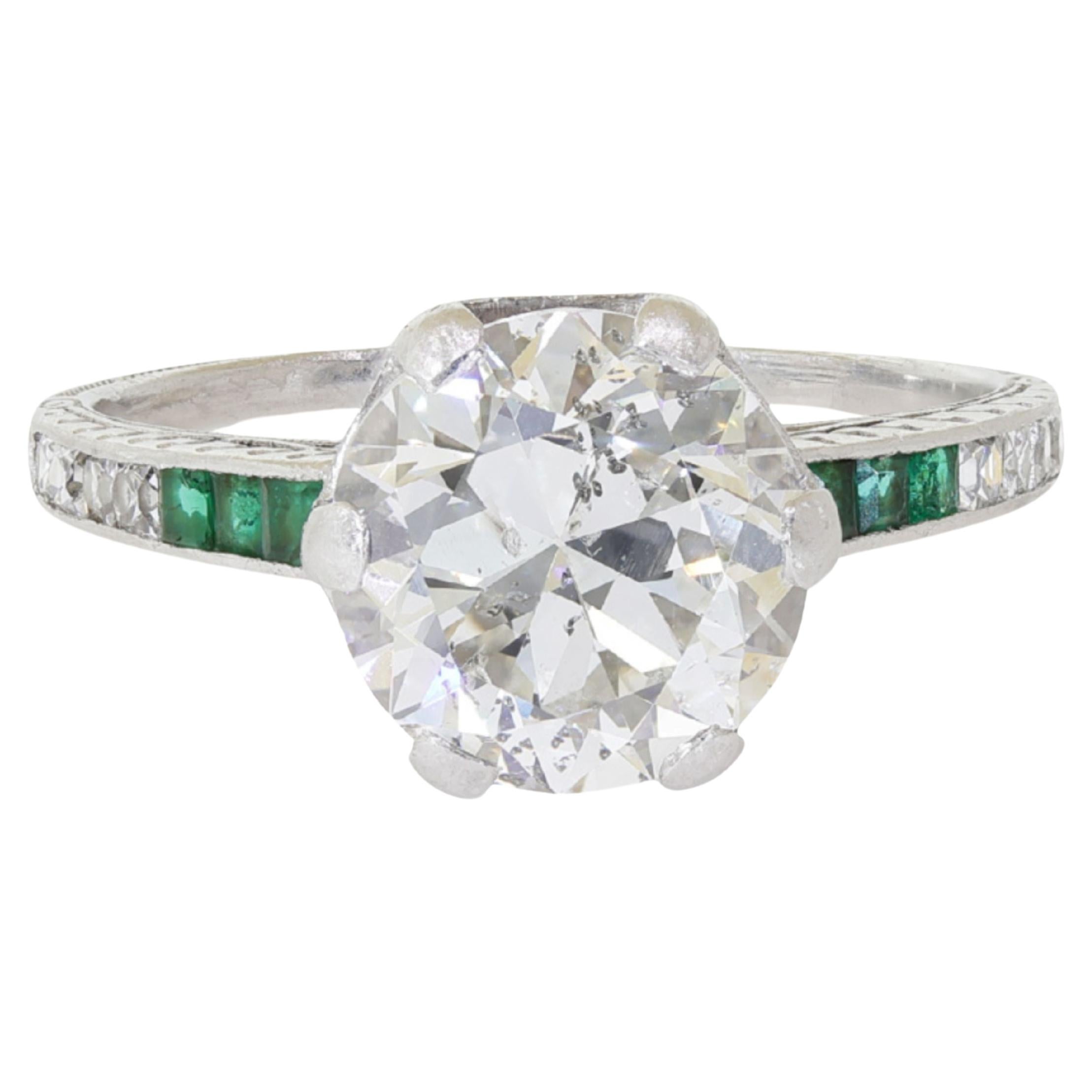 Vintage Art Deco Carved Filigree Platinum Diamond and Emerald Engagement Ring. Featuring a 2.50 carat Old European cut natural diamond center stone. Set in a platinum 6-prong setting that bears a mystical filigree carving that captivates the eye.