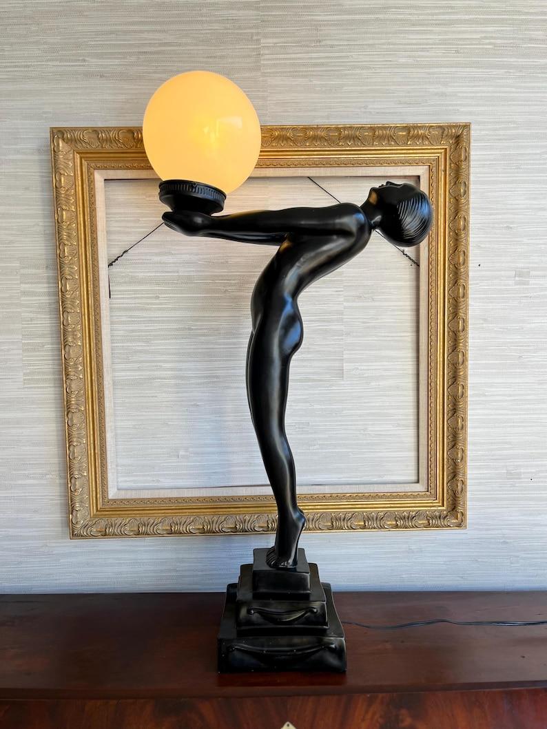 Diese atemberaubende Lampe im Art-Déco-Stil verleiht jedem Raum zeitlose Eleganz. Es zeigt eine anmutige nackte Frau, die auf ihren Zehenspitzen balanciert und sich nach hinten streckt, während sie eine Milchglas-Kugel in den Himmel hebt.

Ihre