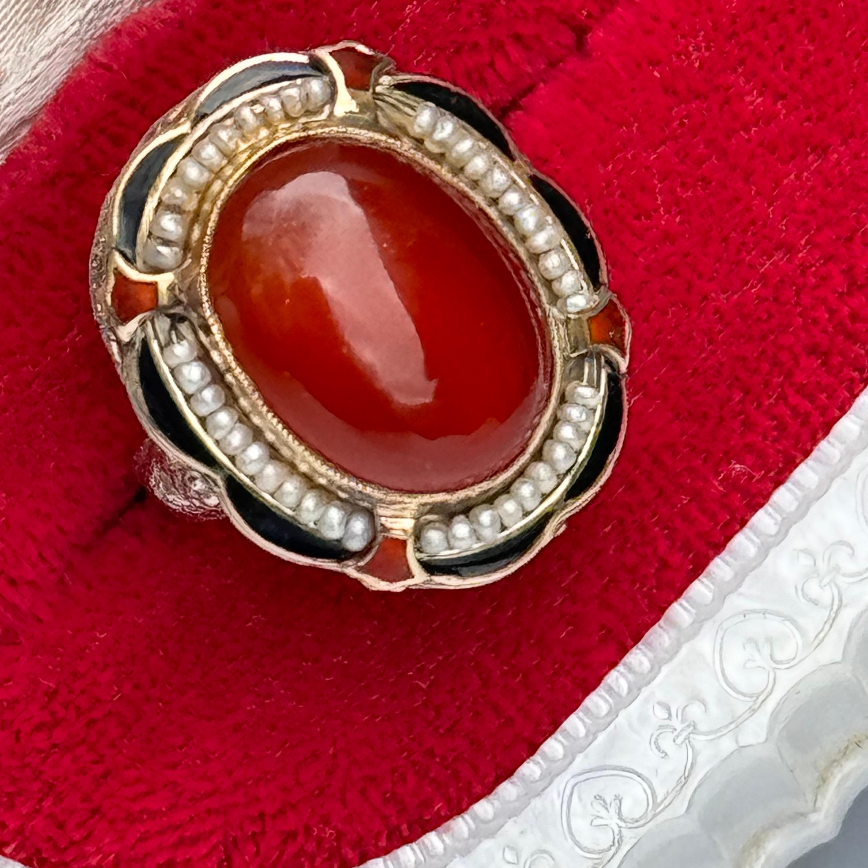 The Art Deco Gold Enamel Ring is a vintage piece featuring intricate filigree work in 14kt white and yellow gold. It showcases a stunning carnelian gemstone accented by delicate seed pearls. Enamel detailing adds vibrant color and depth to the