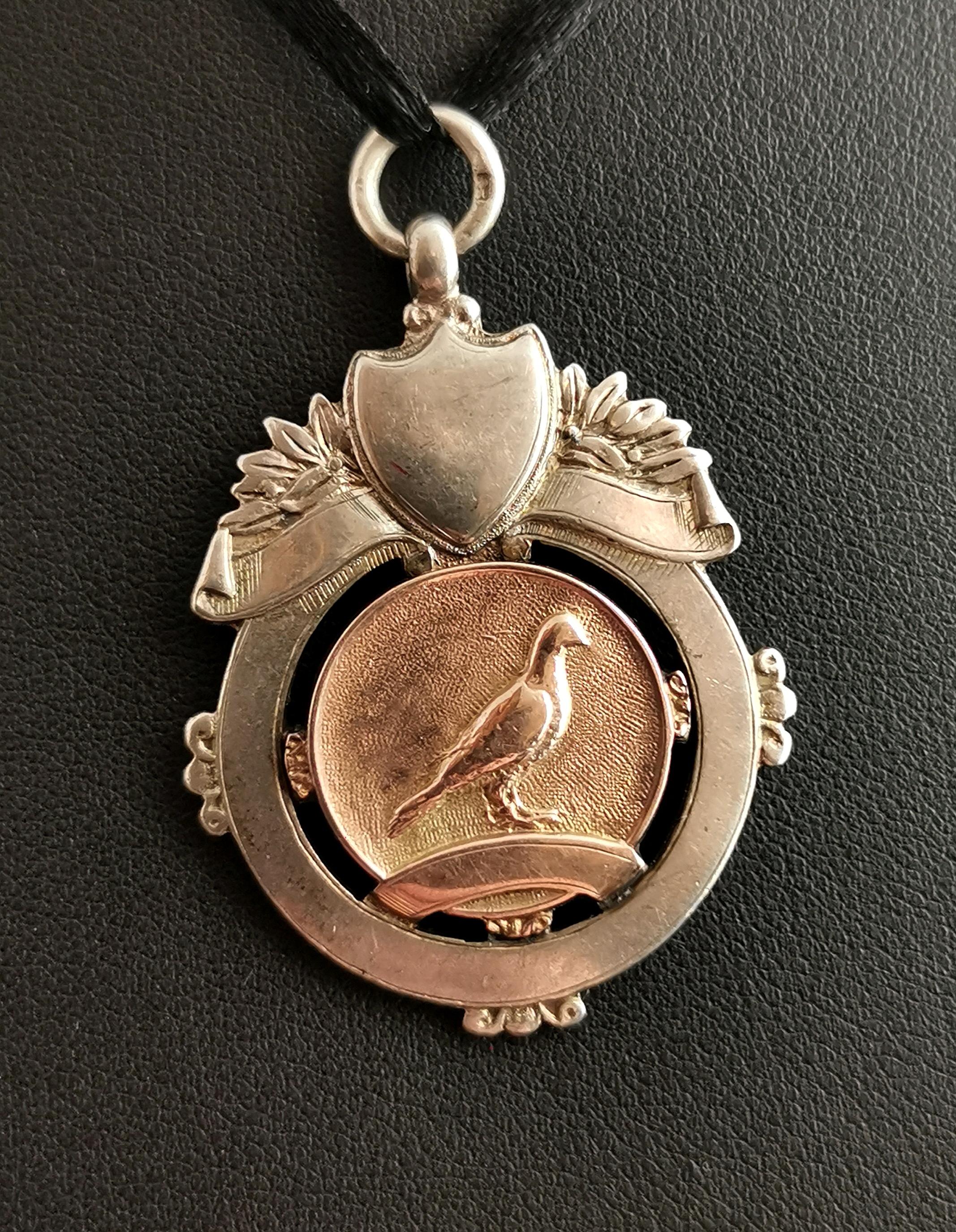An attractive vintage, 1920's sterling silver and Rose gold watch fob pendant.

A nice piece and quite heavy for a fob with a decorative design and a 9kt Rose gold applied plaque to the centre featuring a homing pigeon.

It has a raised shield