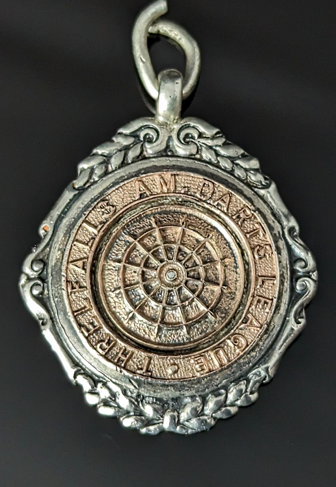 A fantastic vintage Art Deco sterling silver and rose gold watch fob.

It is a circular shaped fob with an applied repousse rose gold dartboard to the front surrounded by scrolls and foliate.

The text around the board reads; Threlfalls AM Darts