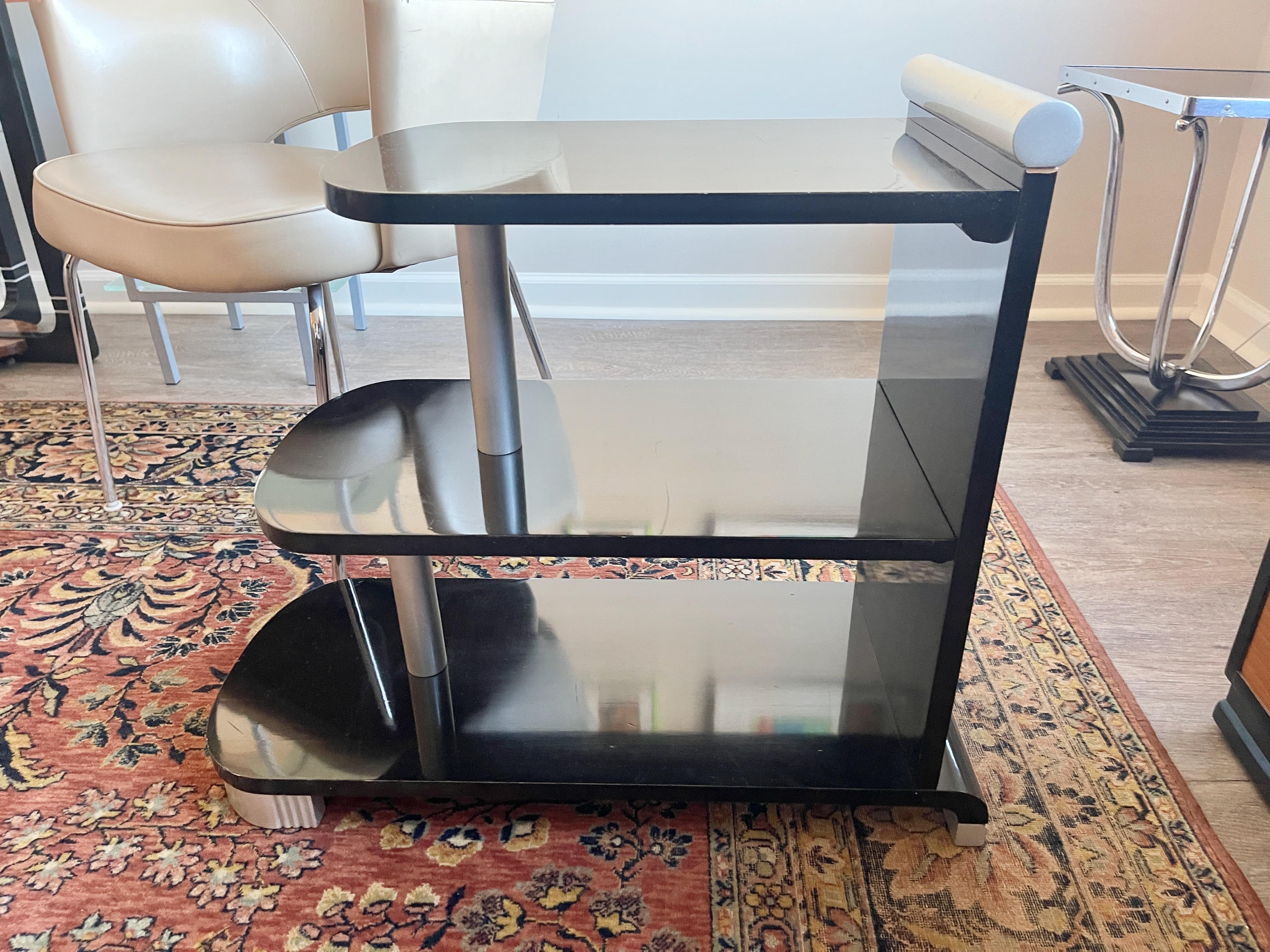 Machine age style vintage three tier table in black lacquer with silver painted accents.

Top shelf 19 inches deep. Bottom shelf 28 inches deep.
9 inches height between shelves.
