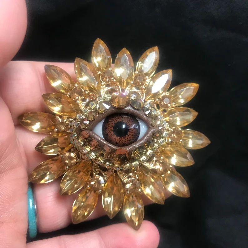 Semplicemente bello! Spilla di dichiarazione in cristallo dorato Art Glass Evil Eye Mid Century Modern con al centro un occhio maligno in vetro artistico. Circondato da cristalli scintillanti dorati Marquise incastonati a mano con un design a