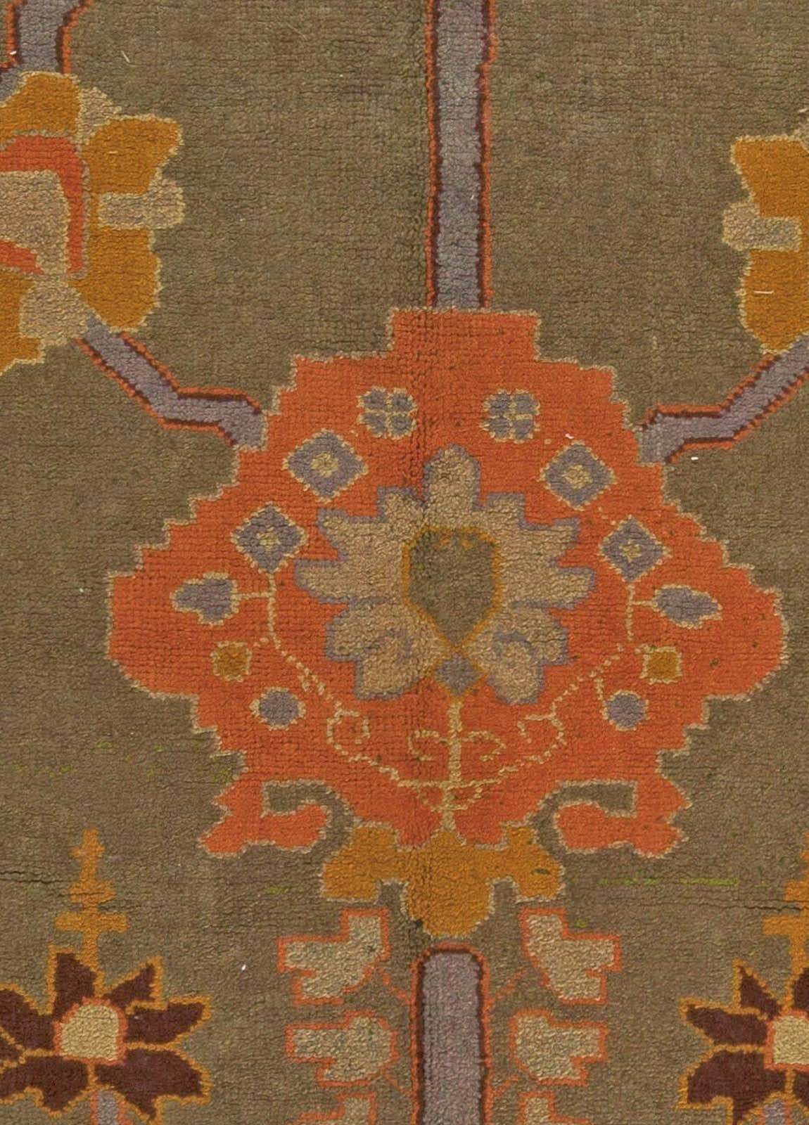 This circa 1920 vintage Arts & Crafts rug by Voysey contains an all-over design of floral motifs in orange, blue and beige against a field of gray-green. A thick orange border featuring botanical abstractions follows the perimeter of the vintage