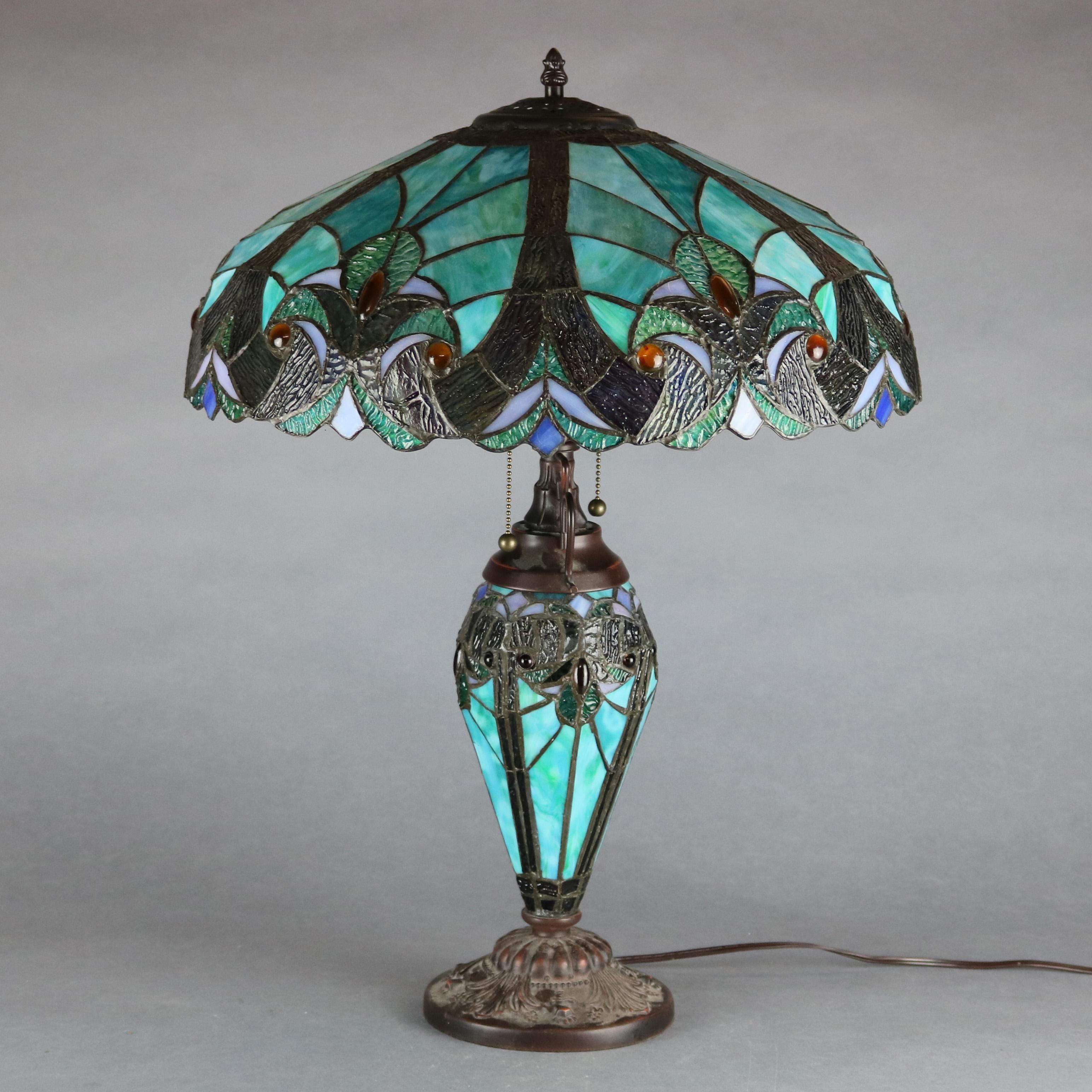 A vintage Arts & Crafts stained glass table lamp in the manner of Tiffany Studios offers mosaic slag and jeweled glass shade in greens and blues with stylized scroll and foliate pattern surmounting matching urn form base having dual independently