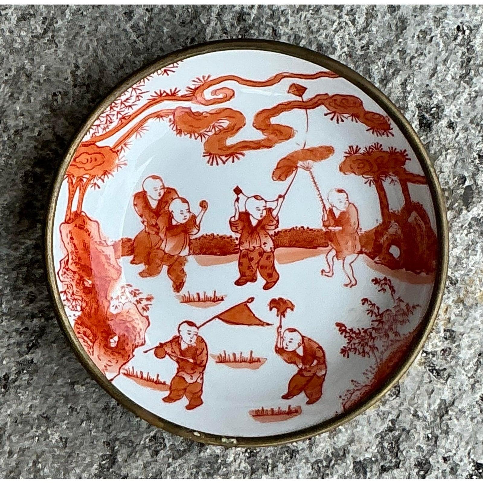 Diese detailreiche und charmante asiatische Schale im Vintage-Stil zeigt ein verspieltes rot-weißes Motiv mit spielenden Kindern, die Drachen steigen lassen und zwischen Bäumen tanzen. Eingerahmt von einem goldfarbenen Metallrand, fängt die Schale