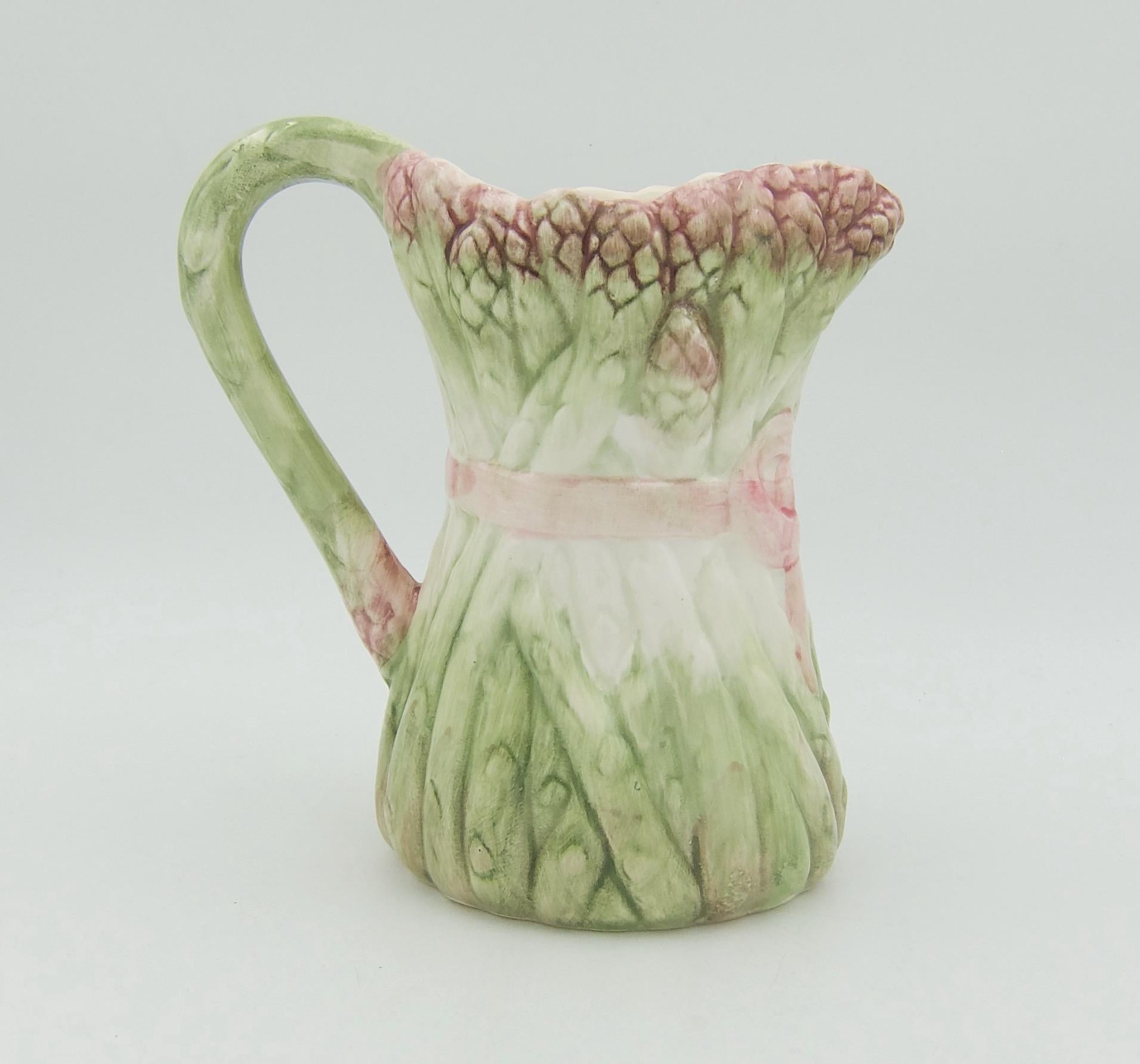 A vintage ceramic pitcher designed to resemble a bundle of green and white asparagus stalks with dark pink tips, tied at the center with a pale pink ribbon and bow. The molded and hand-painted majolica style glazed jug is an ideal size for serving