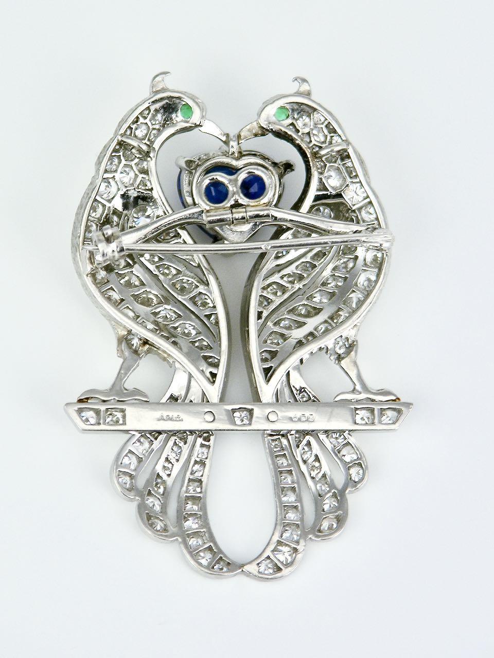 A beautiful Asprey platinum diamond sapphire and emerald peacock brooch consisting of a central heart shaped blue sapphire of 2.75 carats claw set between two stylised birds pave set with 143 diamonds and with emerald eyes with a rollover clasp to