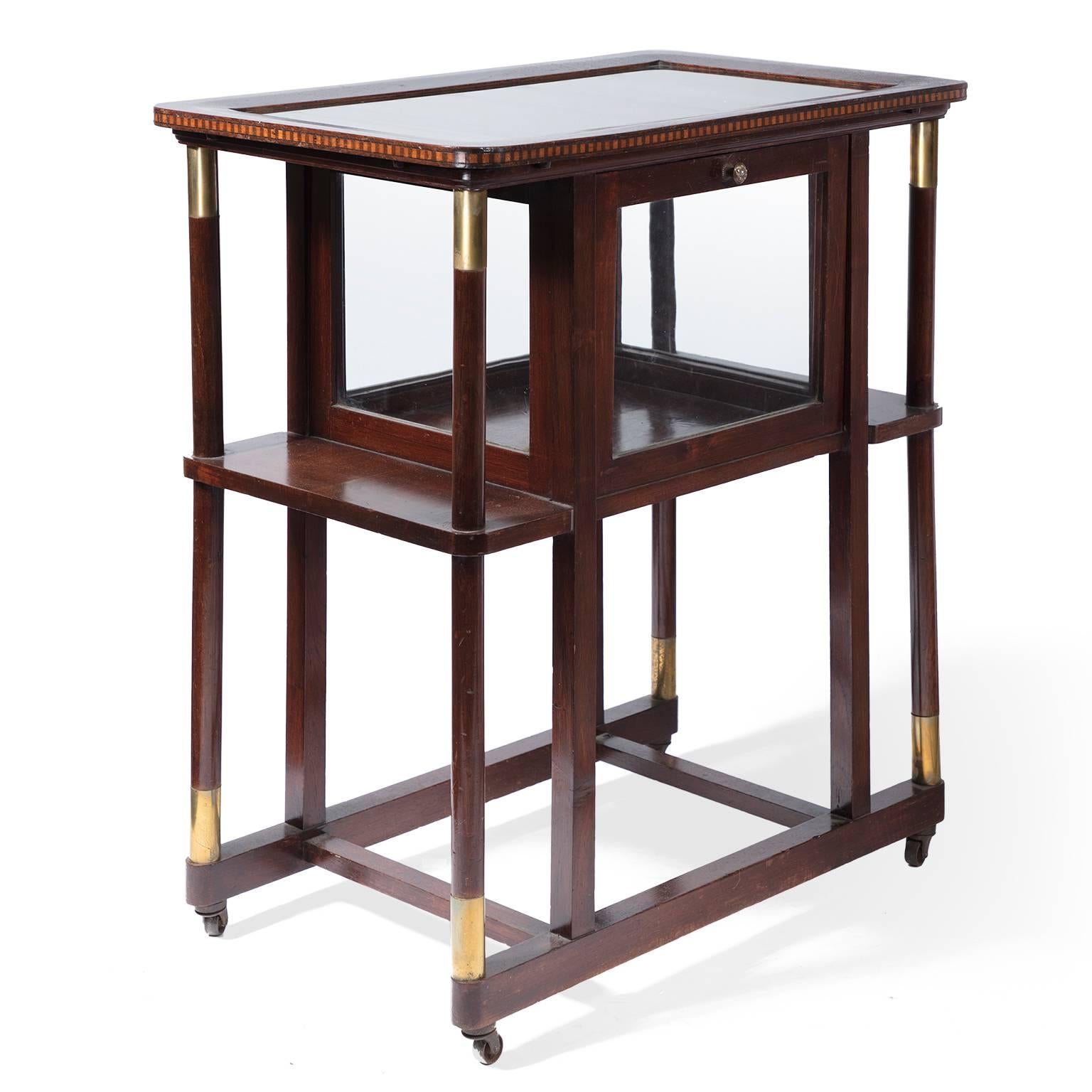 In wood, brass and glass, this early 20th century Austrian bar cart includes two tiers, a removable tray top and hinged glass doors, for easy access to a transparent liquor cabinet. Fine metal details and an inlaid edge add to its bespoke charm,