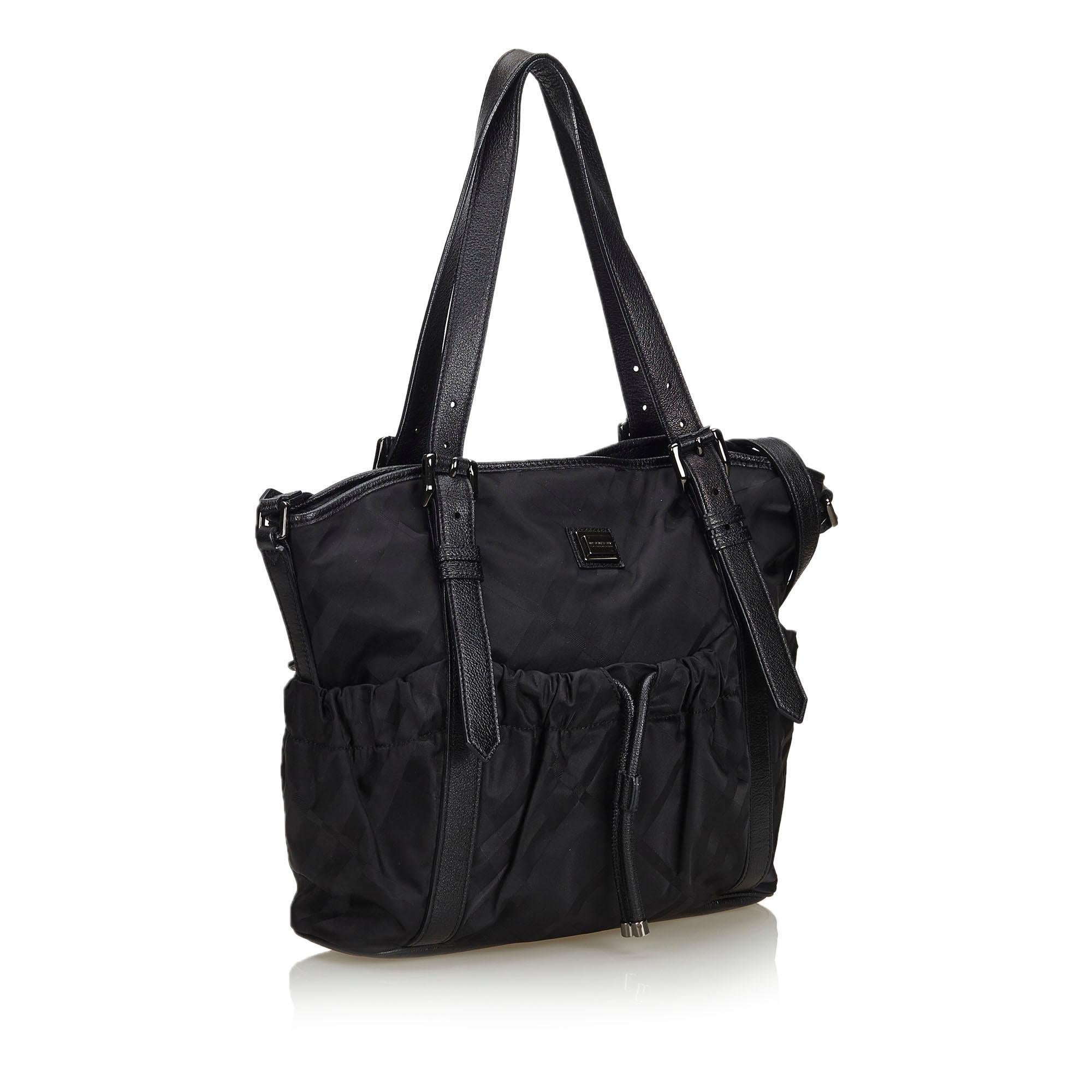 This satchel features a nylon body with leather trim, front and back slip pockets with a drawstring closure, flat leather handles, a flat leather strap, a top zip closure, and interior zip and slip pockets. It carries as B+ condition