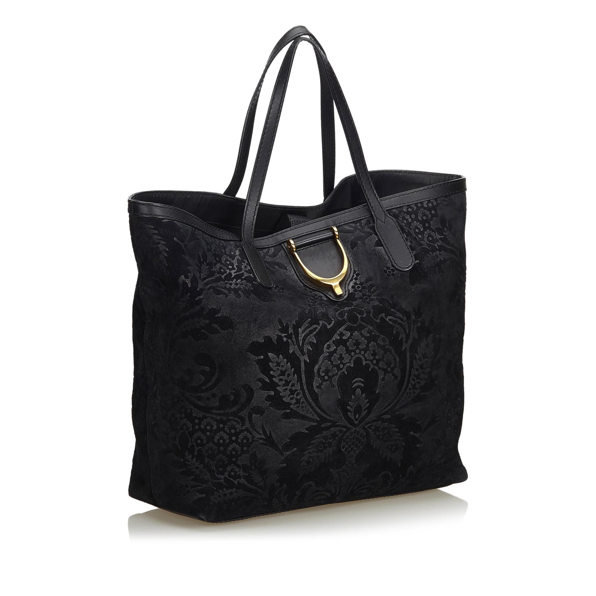 The Stirrup tote bag features a suede brocade body with gold-tone hardware, flat leather straps, an open top with magnetic snap button closure, and interior zip and slip pockets. It carries as B+ condition rating.

Inclusions: 
Dust