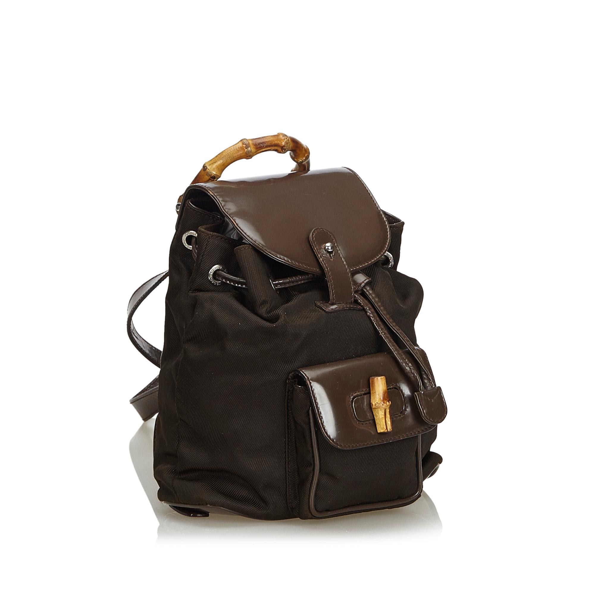 This backpack features a nylon body with leather trim, an exterior front flap pocket with bamboo twist lock closure, flat leather back straps, a bamboo top handle, top flap with buckle closure, drawstring closure, and an interior zip pocket. It