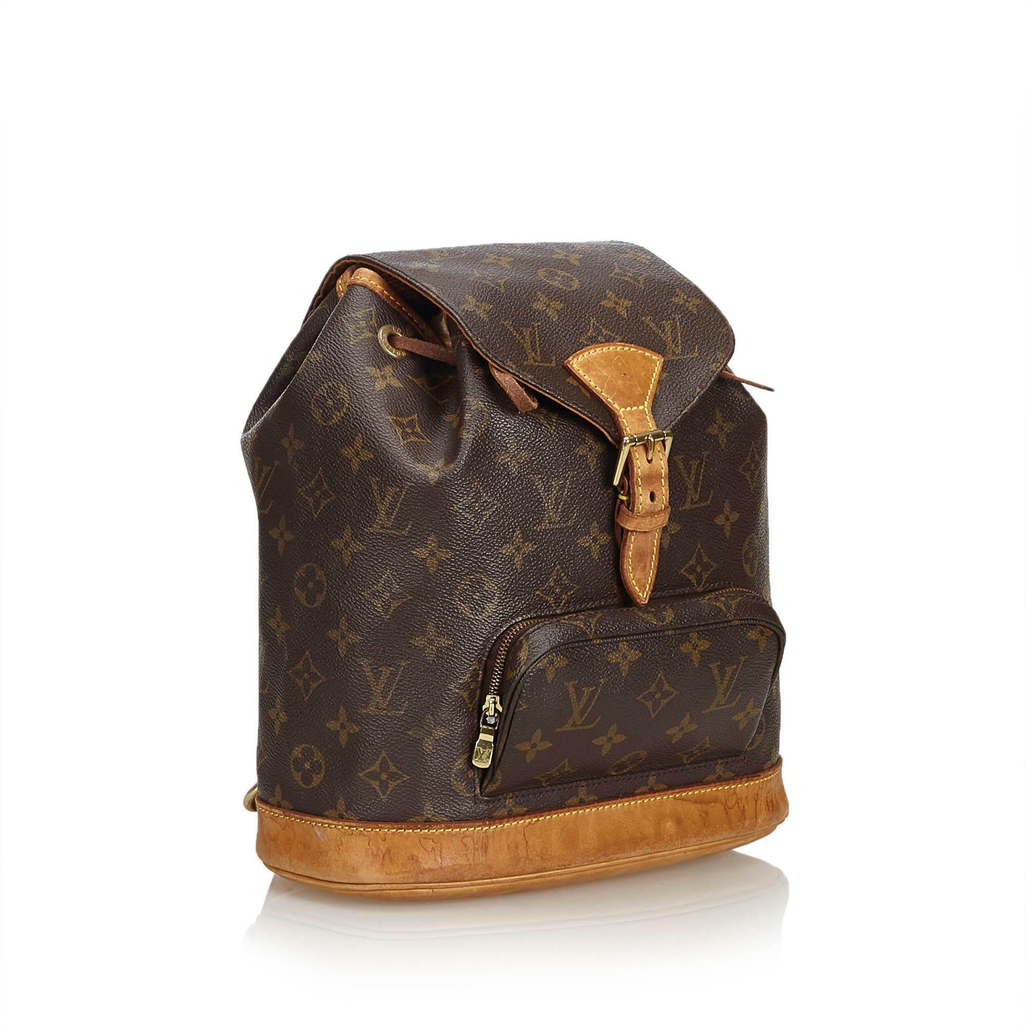 The Montsouris MM features a monogram canvas body, flat shoulder straps, a leather bottom, a front flap with a belt buckle detail and a magnetic closure, a top drawstring closure, and an interior slip pocket. It carries as B condition