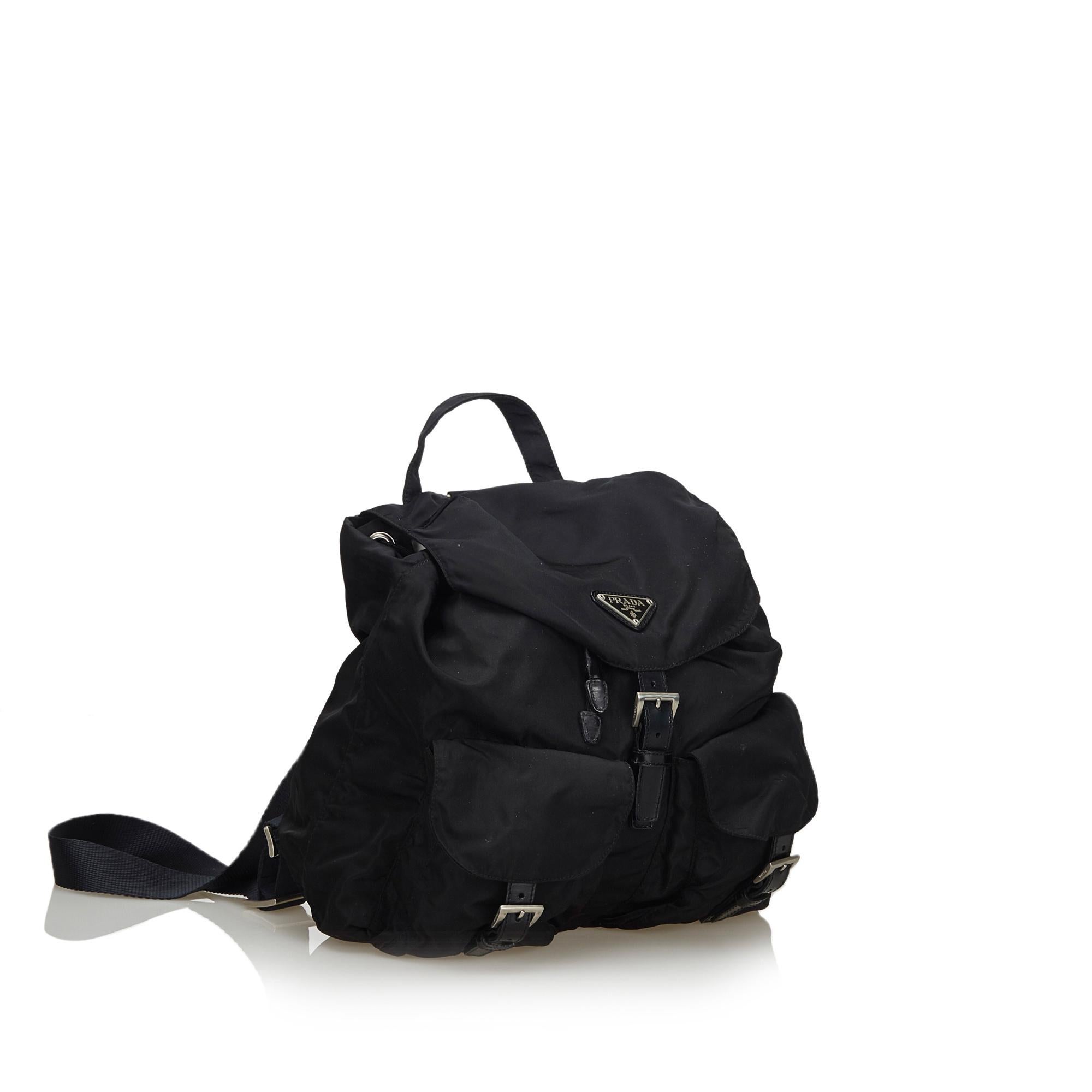 This backpack features a nylon body with leather trim, front exterior flap pockets with leather buckle closure, flat back straps, a flat top handle, a top flap with a drawstring closure, and an interior zip pocket. It carries as B+ condition