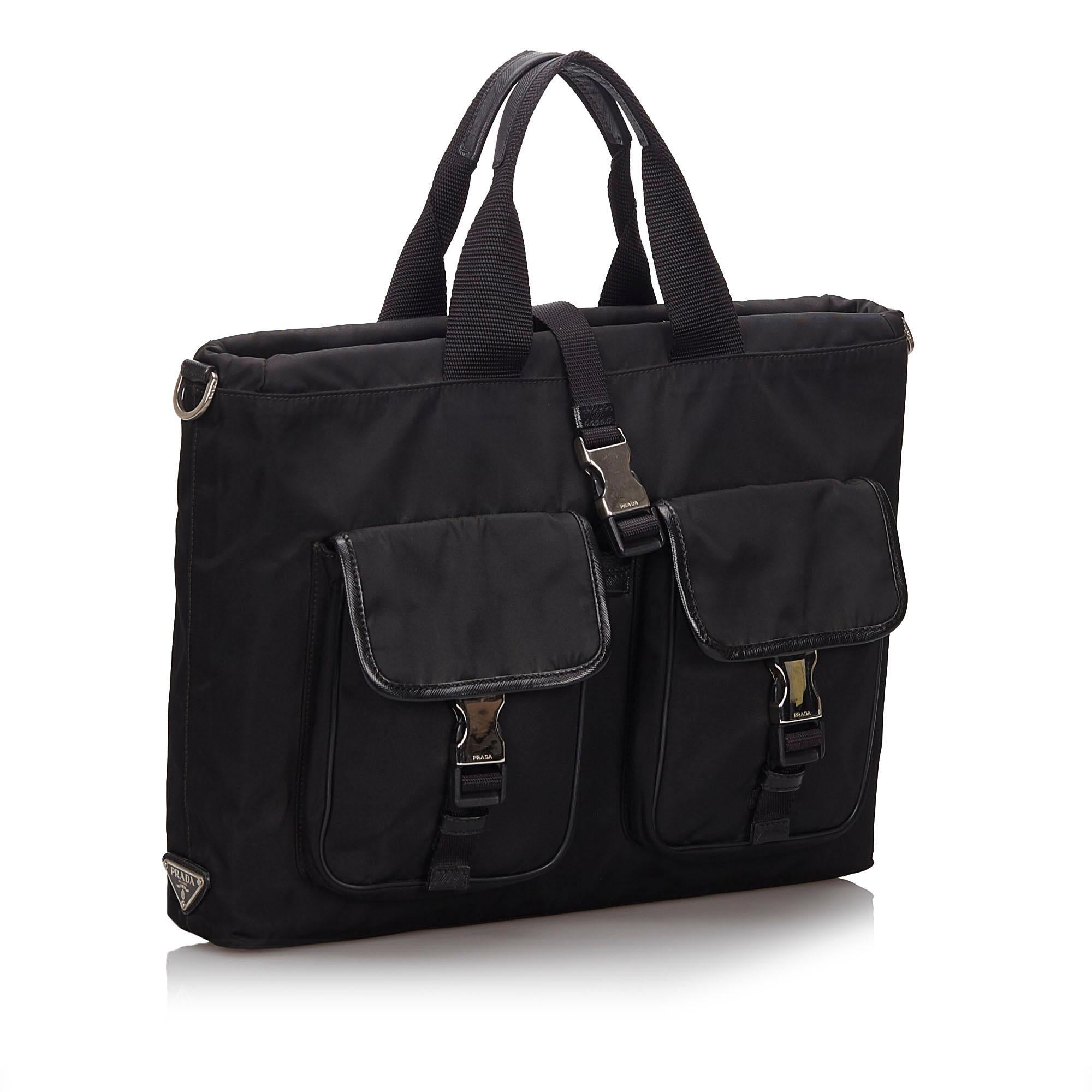 This satchel features a nylon body with leather trim, front exterior flap pockets with buckle closures, a back exterior zip pocket, flat handles, a detachable flat strap, a top strap with a buckle closure, and an open top. It carries as B+ condition