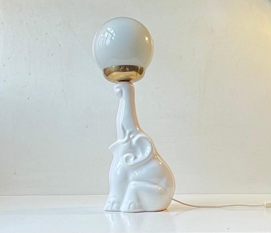 Vintage table lamp in the shape of a baby elephant in a cute balancing act with a 'ball'. Its made from white glazed porcelain featuring an opaline glass shade with brass rest/socket. We suspect it might have been made by Lladró in Spain during the