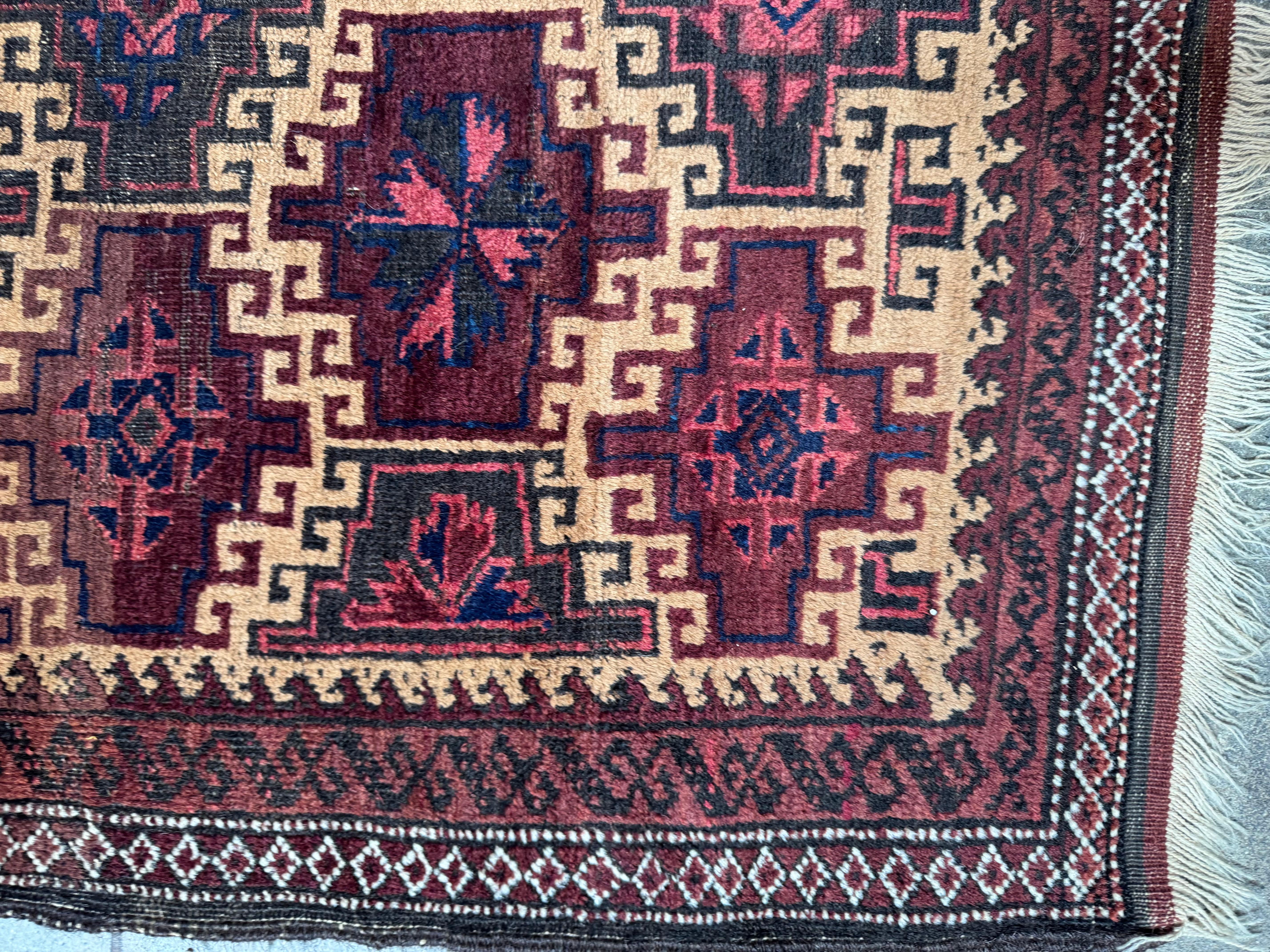 Dieser auffällige Vintage-Teppich von Balutch zeichnet sich durch ein kühnes, geometrisches Layout mit sich wiederholenden Stammes-Sternmedaillons in tiefem Burgunderrot, sattem Schwarz und Akzenten in Marine und Elfenbein aus. Die komplizierten