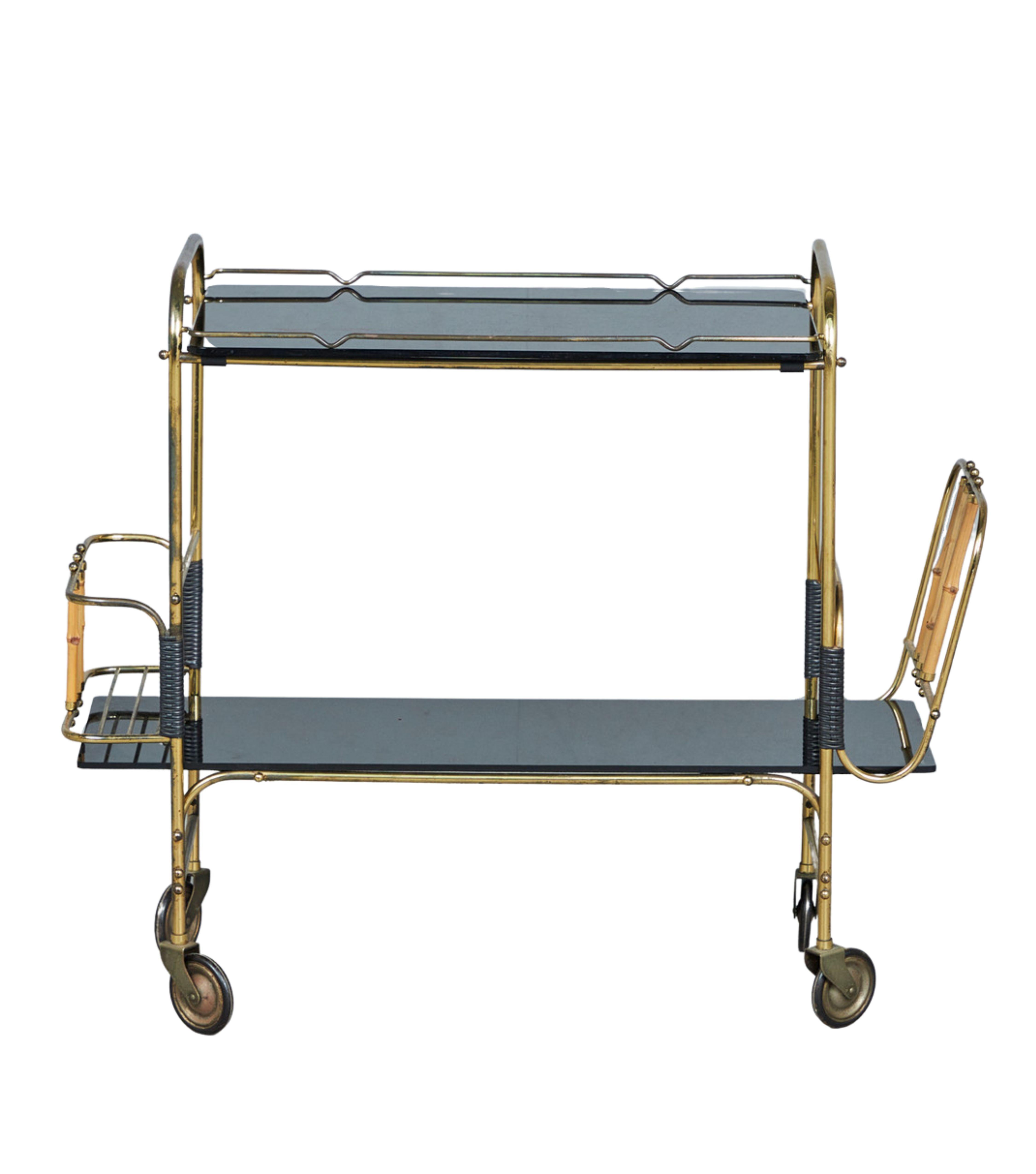 This German bar serving trolley from the 1950s made of metal coated in brass, with a unique design that includes bamboo cane braces. The trolley features two shelves made of black glass and is equipped with four wheels, a newspaper stand, and a