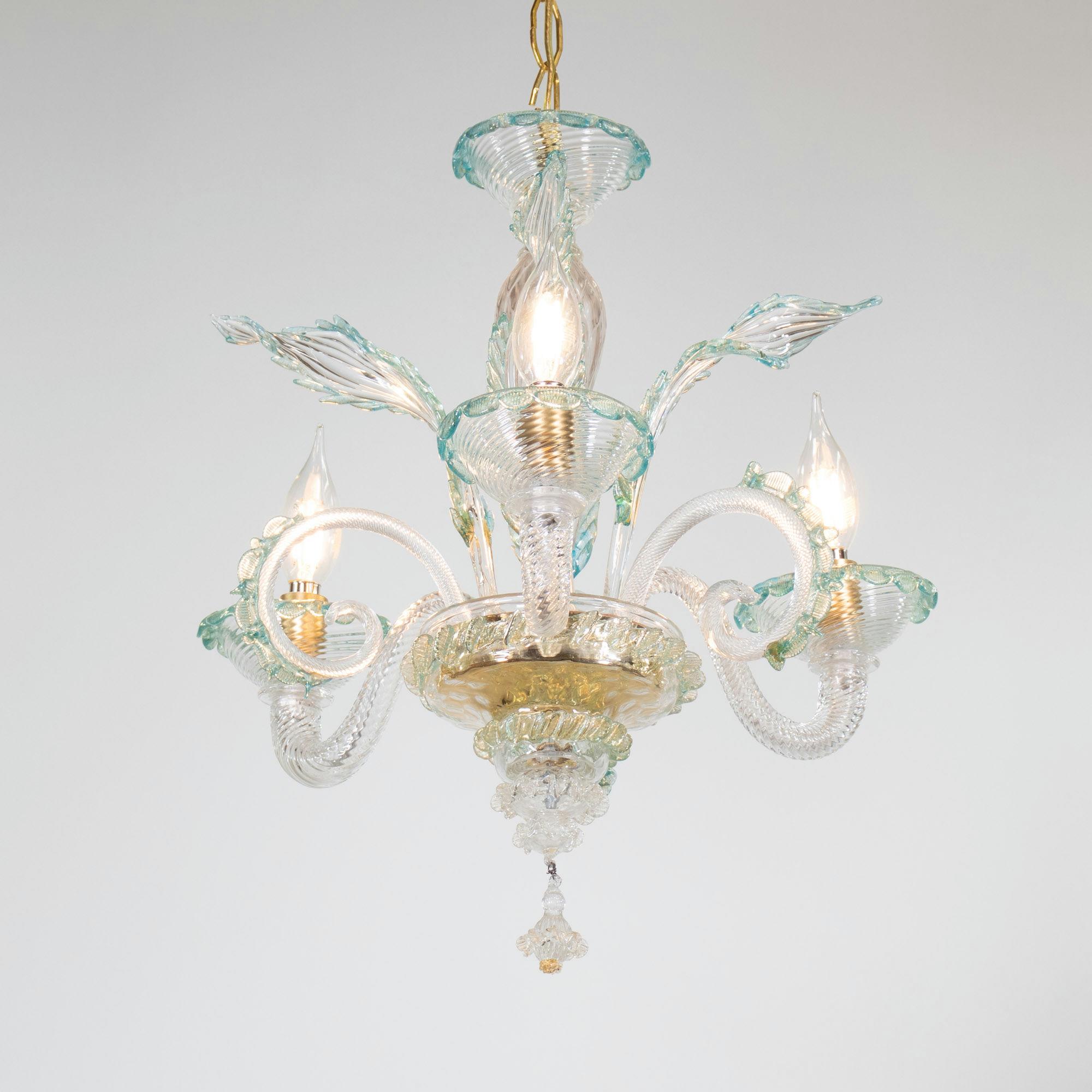 Questo lampadario artigianale in cristallo di Murano ha tre bracci a voluta e presenta una colonna centrale a bulbo con dettagli naturali che spuntano, qui stilizzati con foglie e ornamenti a voluta. Il corpo e i bracci sono realizzati in vetro