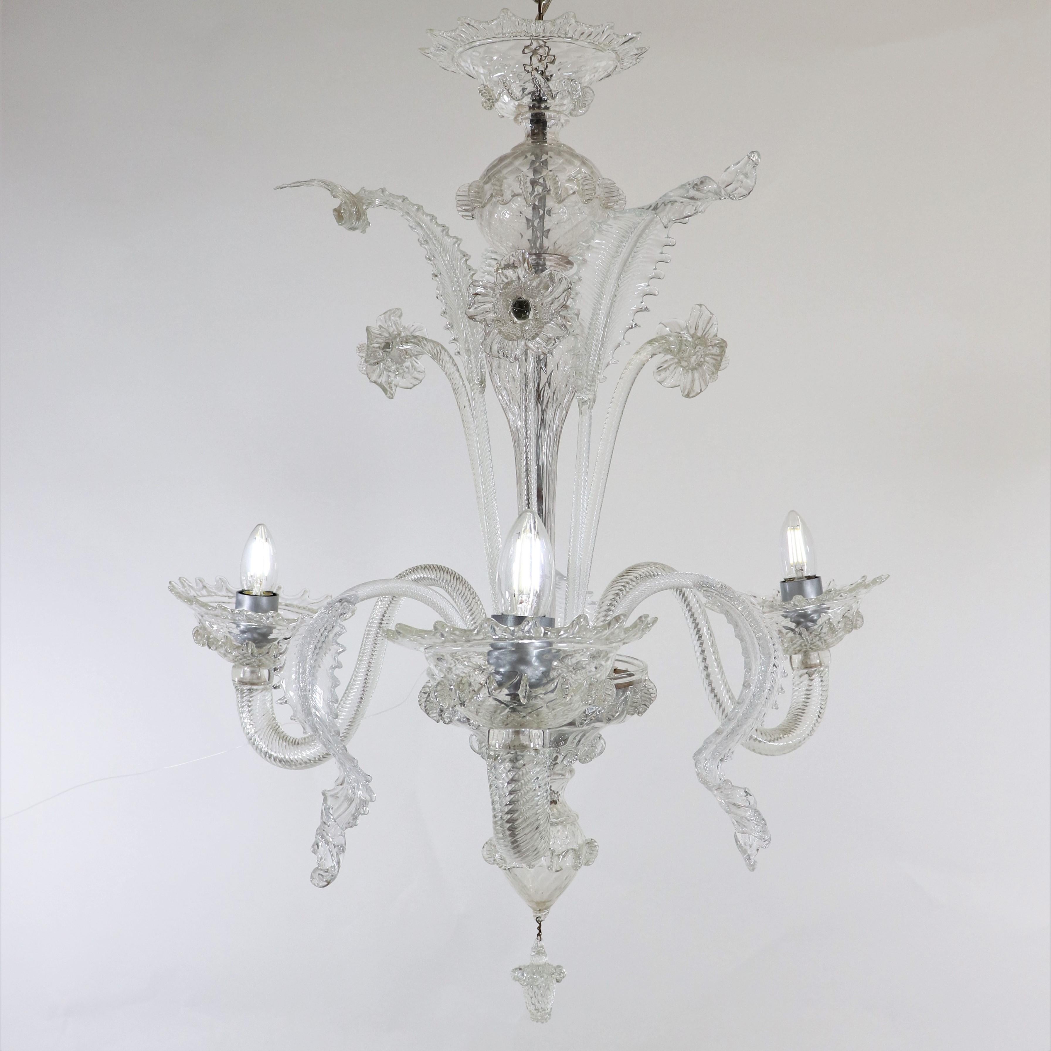 Questo lampadario artigianale in cristallo di Murano ha tre bracci a voluta e presenta una colonna centrale a bulbo con dettagli naturali che spuntano, qui stilizzati con narcisi e foglie. Il corpo e i bracci di questo pezzo sono realizzati in vetro