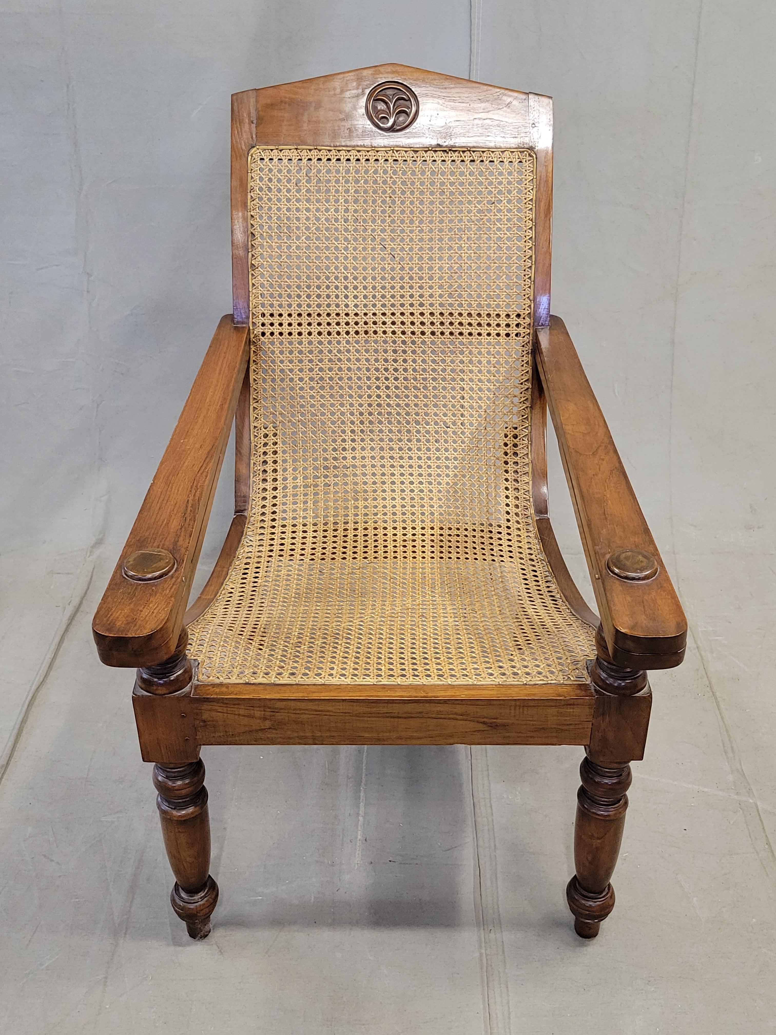 A classic vintage 1990s teak plantation lounge chair with a taught hand-caned seat and backrest. This chair is unique with a handcarved leaf medallion on the headrest and large buttons on the armrests. As with this style of plantation chair,