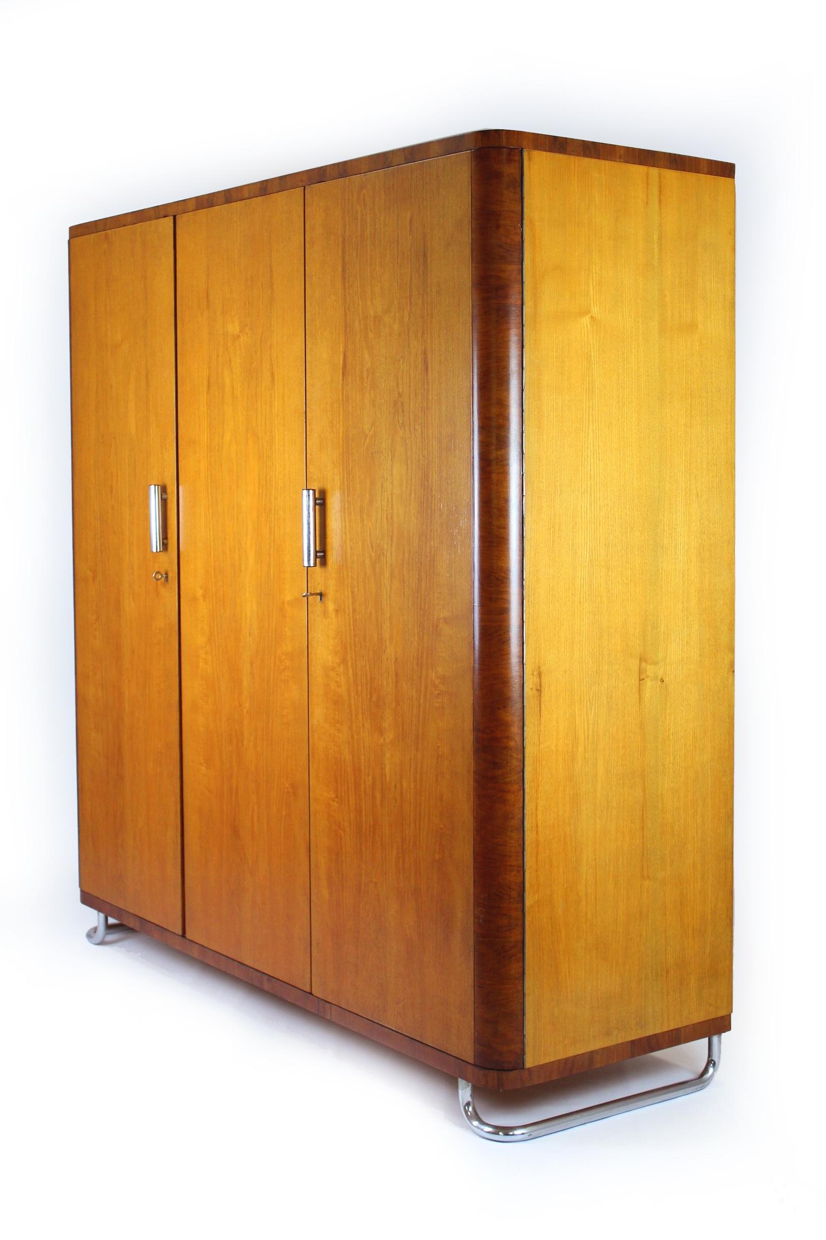 Cette armoire de style Bauhaus a été produite en Tchécoslovaquie dans les années 1930. Il comporte trois portes, une tringle à linge interne et cinq étagères. L'armoire repose sur des pieds chromés et possède des poignées cylindriques chromées. Très