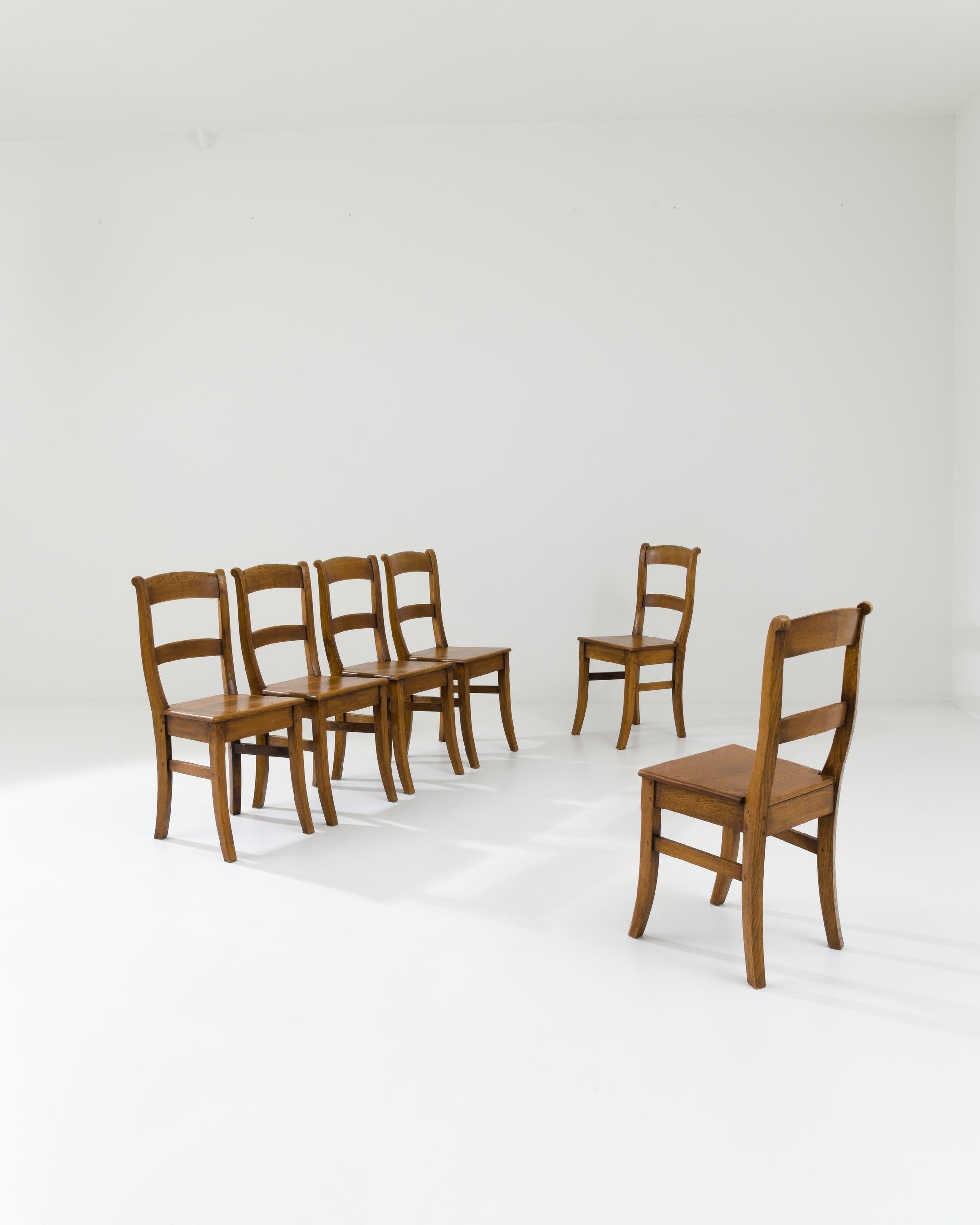 A set of wooden dining chairs made in 20th century Belgium. With swooping seat backs and gracefully bent legs, these chairs exude the grace of simplicity. The high-backed form gives these chairs a scholarly form, the rich brown seats, polished to a