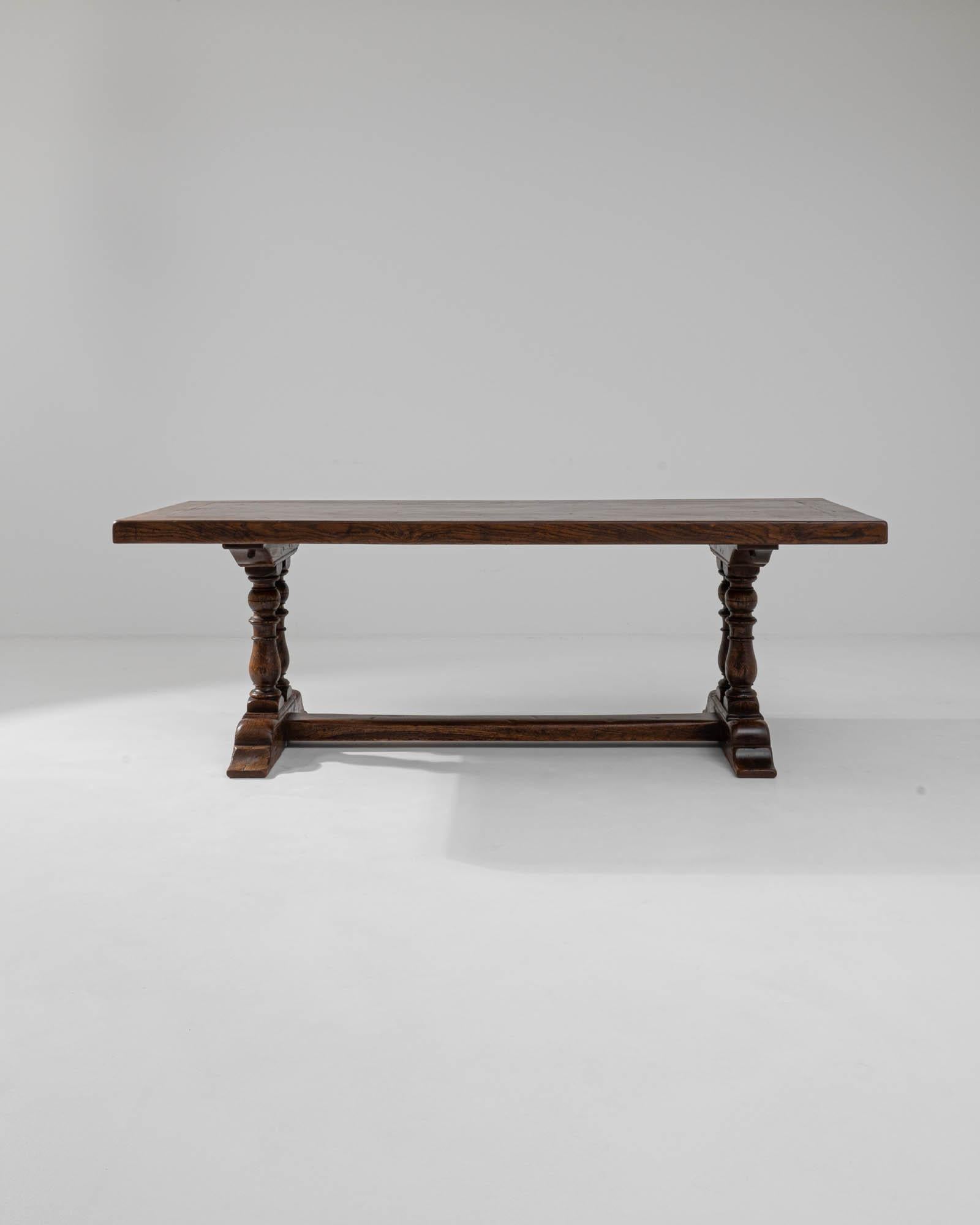 A wooden dining table made in 20th century Belgium. Two sets of lavishly legs support each end of this ornate table, connected by a long stretcher in a trestle fashion. The gleaming, rich oak shines with remarkable luster. Exuding an elegant, yet