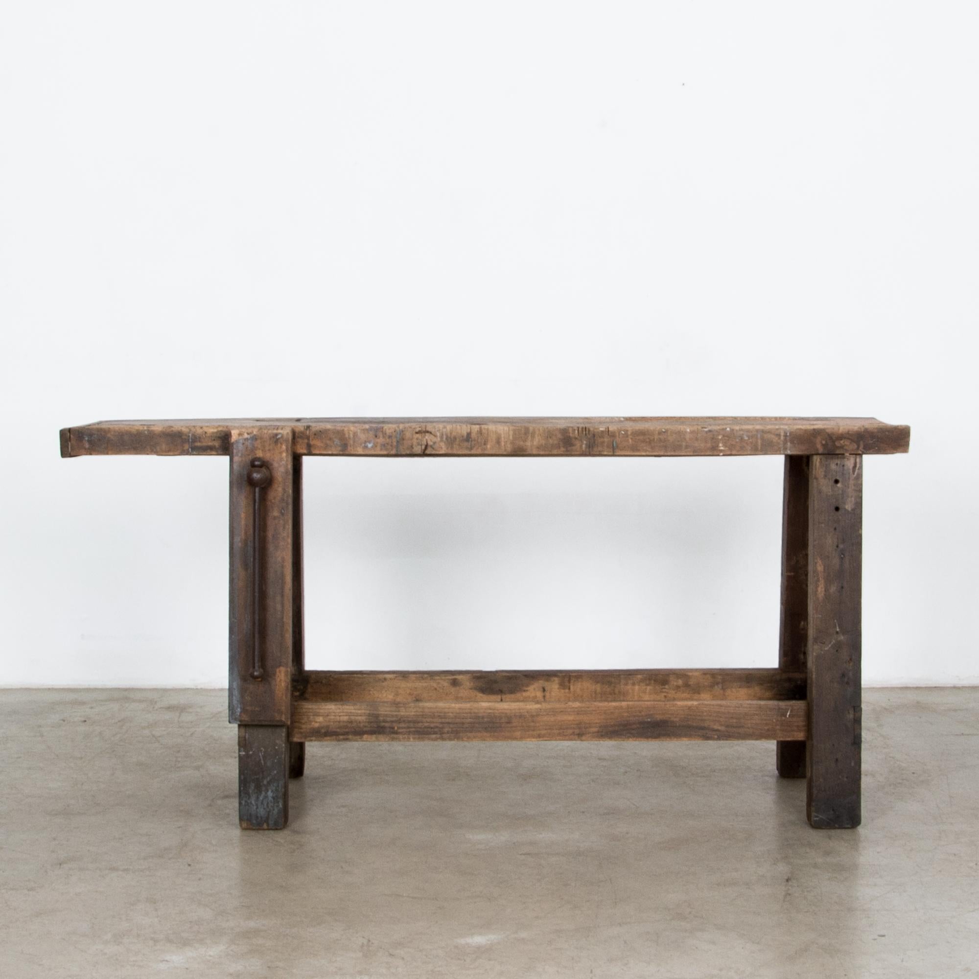 This vintage wooden work table was made in Belgium in the 1950s. A long tabletop is supported by sturdy trestle legs, one of which features a bench clamp. The tabletop is extended on one side, providing unfettered workspace. The dark wood is