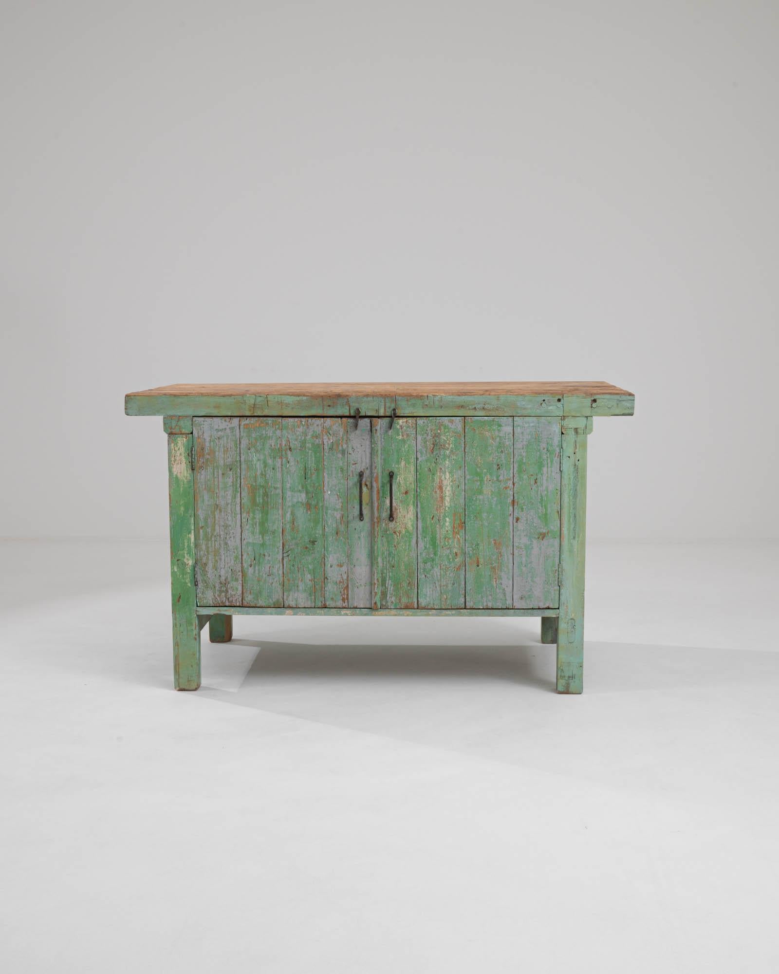 A wooden work table made in 20th century Belgium. The mint green coat of paint has faded to expose underlying browns and beiges along the exterior of this mysterious cabinet. Inside, two drawers perch upon its upper left side, speckled with frosty