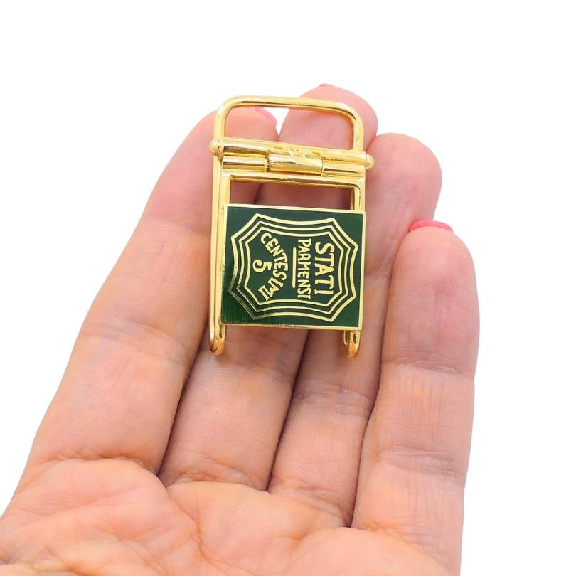This vintage 14k gold pendant plays with the idea of utility and nostalgia in a really clever way. Shaped like a miniature belt buckle, it features a hinged mechanism at the top and a green enamel plate on the front. The inscription reads 