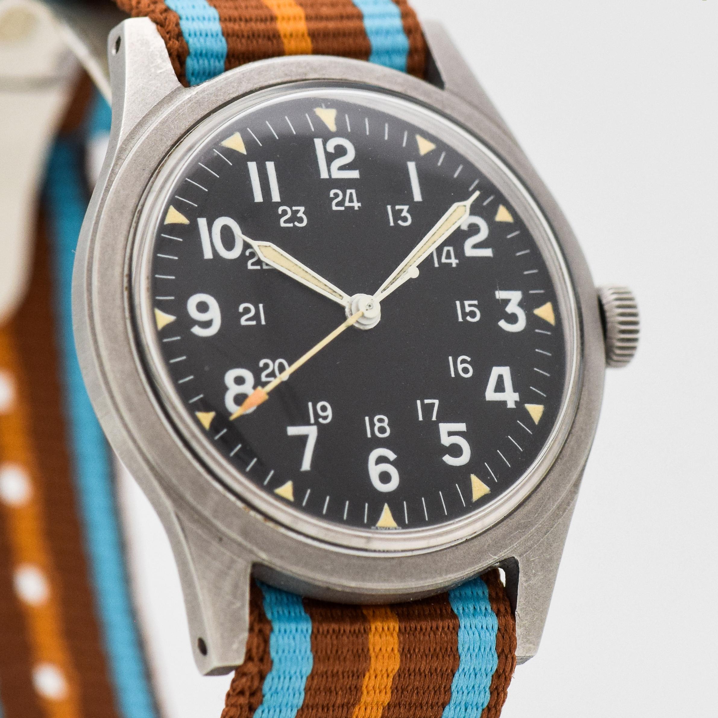 1967 Vintage Benrus Military Stainless Steel watch with Original Black Dial with White Luminous Arabic Numbers. 34mm x 40mm lug to lug (1.34 in. x 1.57 in.) - 17 jewel, manual caliber movement. Equipped with a Nylon-style Brown, Blue &