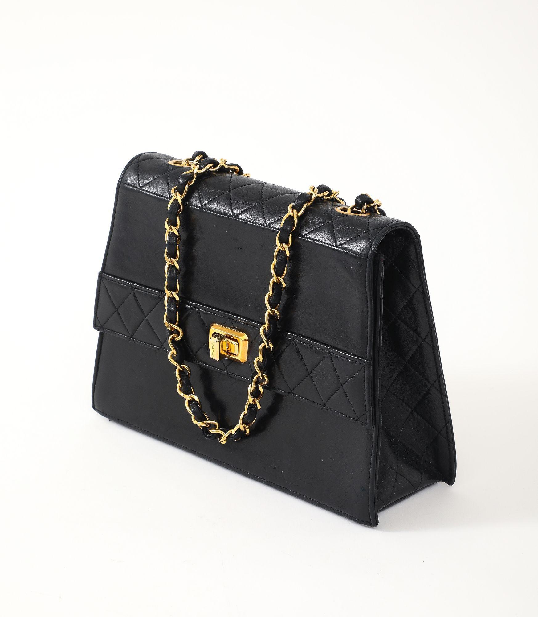 A vintage black Chanel handbag with a flap-style closure, featuring a signature diamond-quilted leather body and a shoulder strap composed of a thick, gold-tone chain interwoven with black leather. The flap is secured with a prominent gold-tone CC
