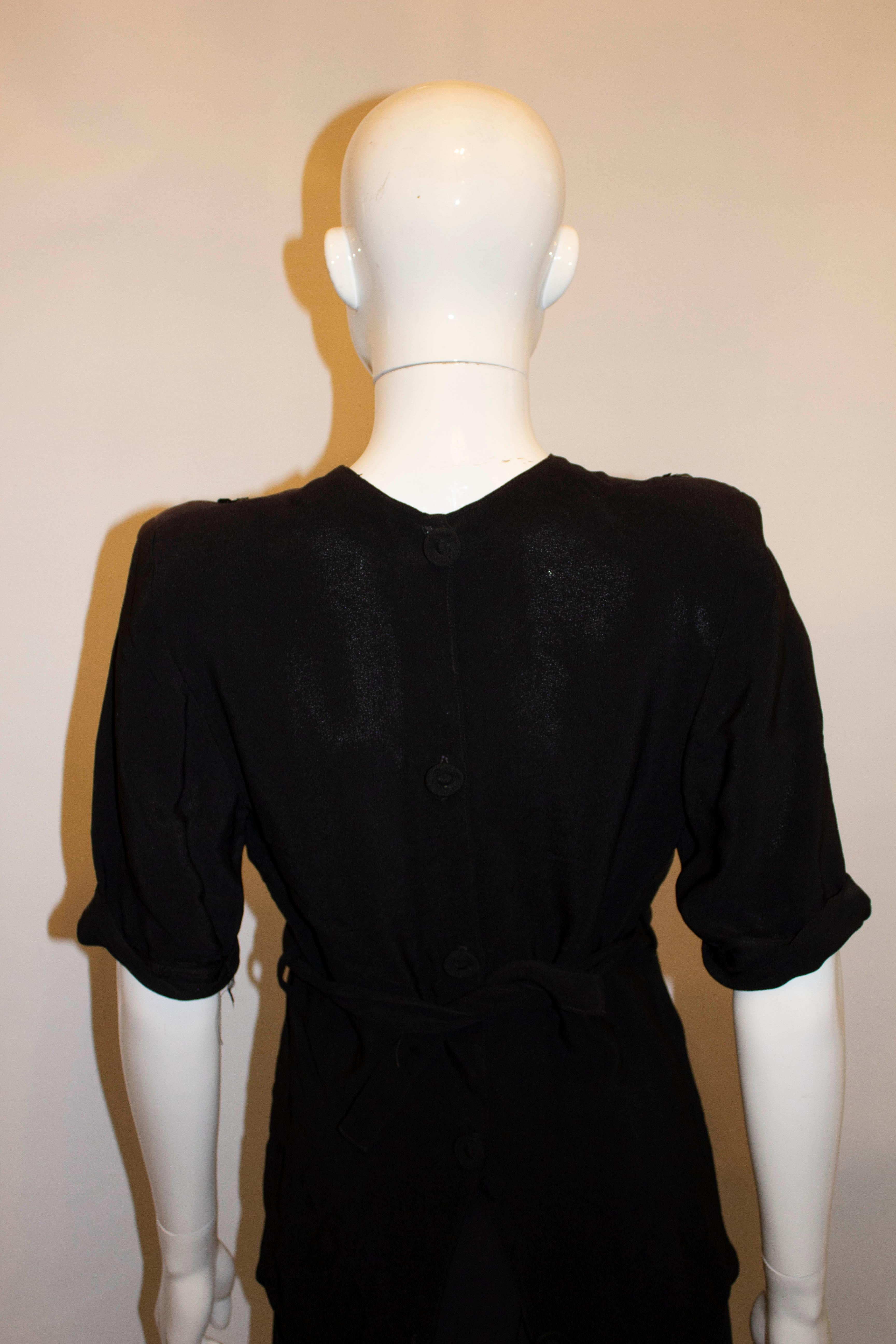 Ein wunderschönes schwarzes Abendkleid aus Krepp aus den 1940er Jahren. Das Oberteil hat Falten  und Pailletten-Detail auf der Vorderseite, stoffbezogene Knopföffnung auf der Rückseite.  und einem Stoffgürtel mit Zopfmuster. An beiden Schultern gibt