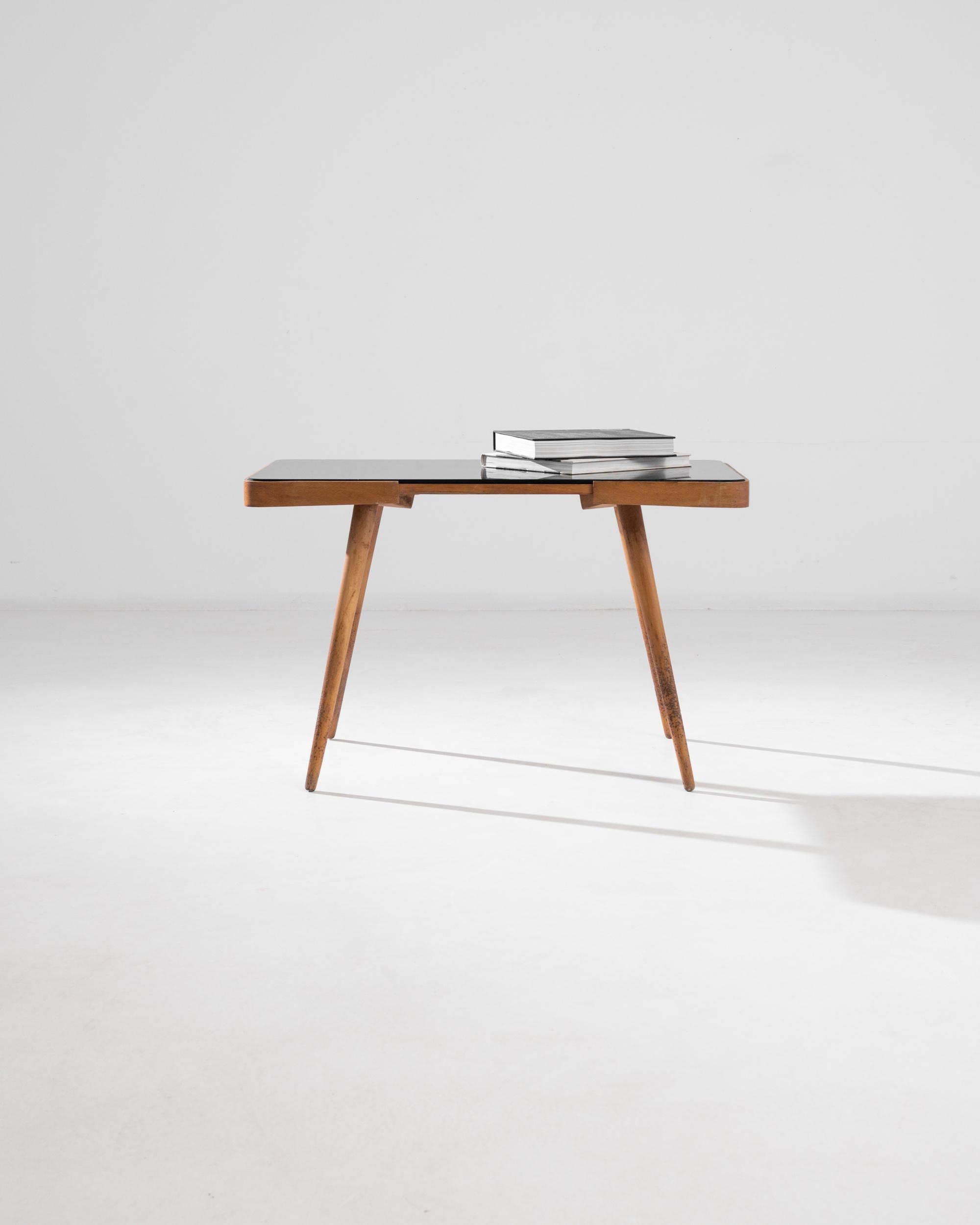 A 20th century wooden table by renowned Czech designer Jirí Jiroutek, this stylized coffee table embodies the mid-century elegance of Czech furniture design. An immaculate black glass lays on a wooden top panel, balanced upon slender splayed legs.