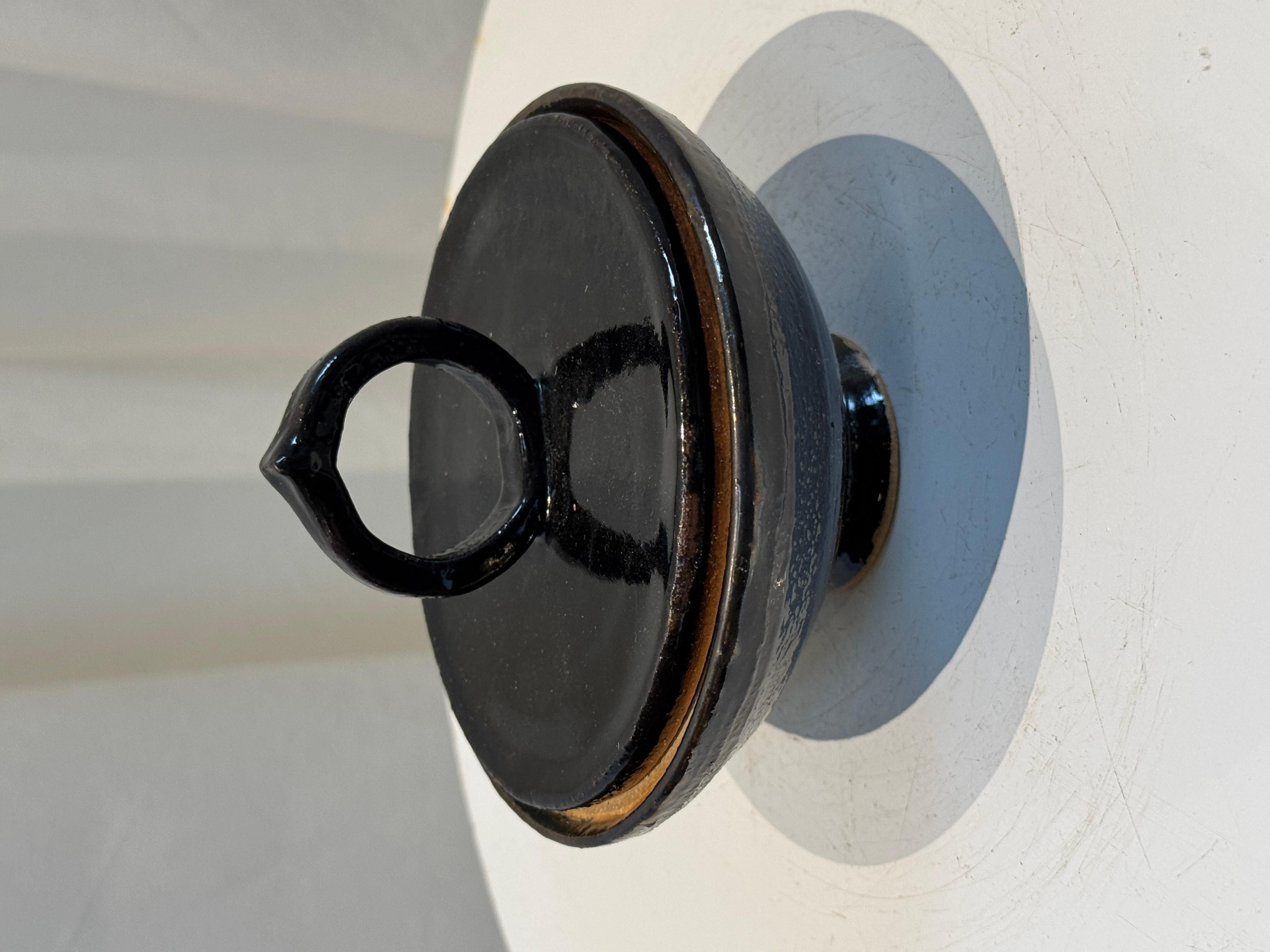 Este llamativo cuenco vintage de cerámica con tapa presenta un profundo y brillante esmalte negro, que contrasta maravillosamente con sus detalles de arcilla natural. El asa en forma de anillo, elegantemente modelada a mano en la parte superior de