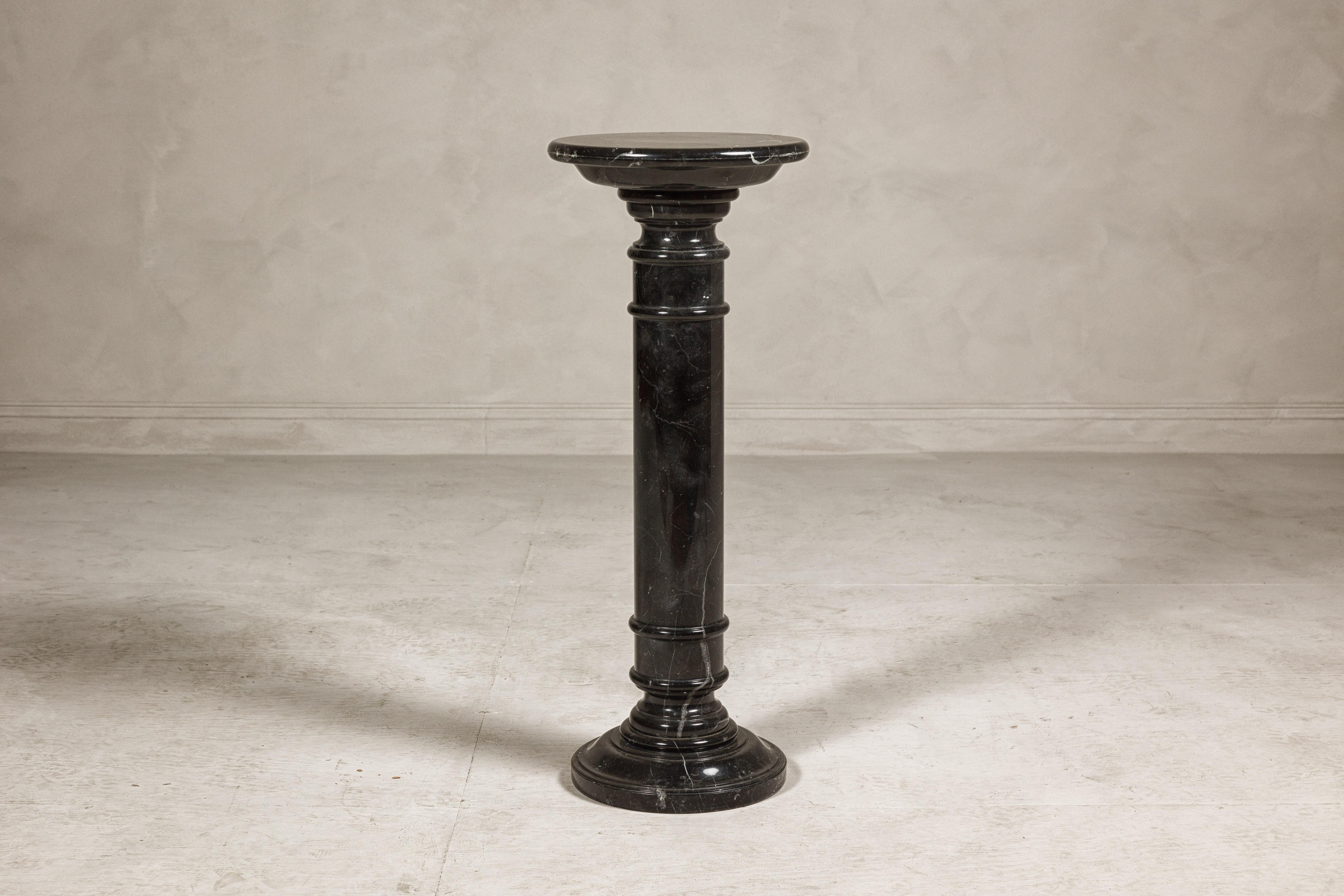 A vintage black marble column shaped pedestal with white veining and stepped base. This vintage black marble column-shaped pedestal is a striking piece that combines the grandeur of classical design with the sleek elegance of modern aesthetics. The