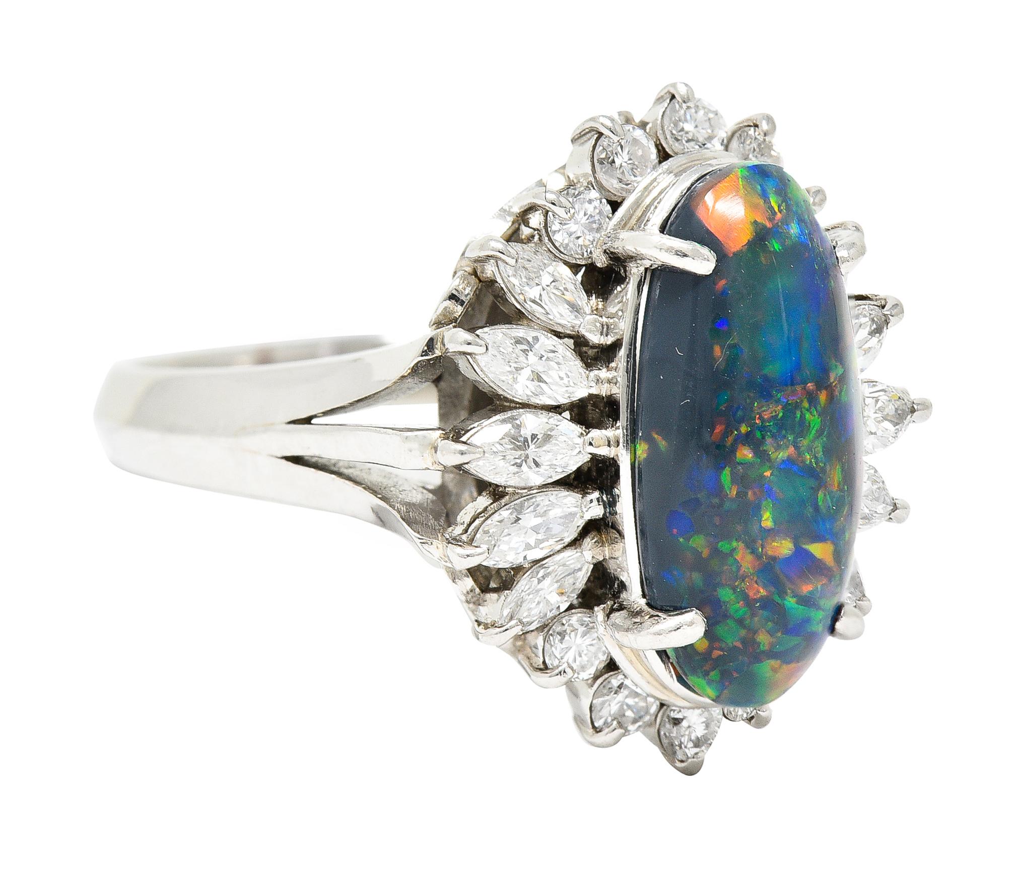Centering an oval shaped black opal cabochon measuring 6.5 x 13.5 mm. Semi-translucent black in body color with spectral play-of-color. Prong set with a halo surround of marquise cut diamonds. With additional round brilliant cut diamonds - all prong