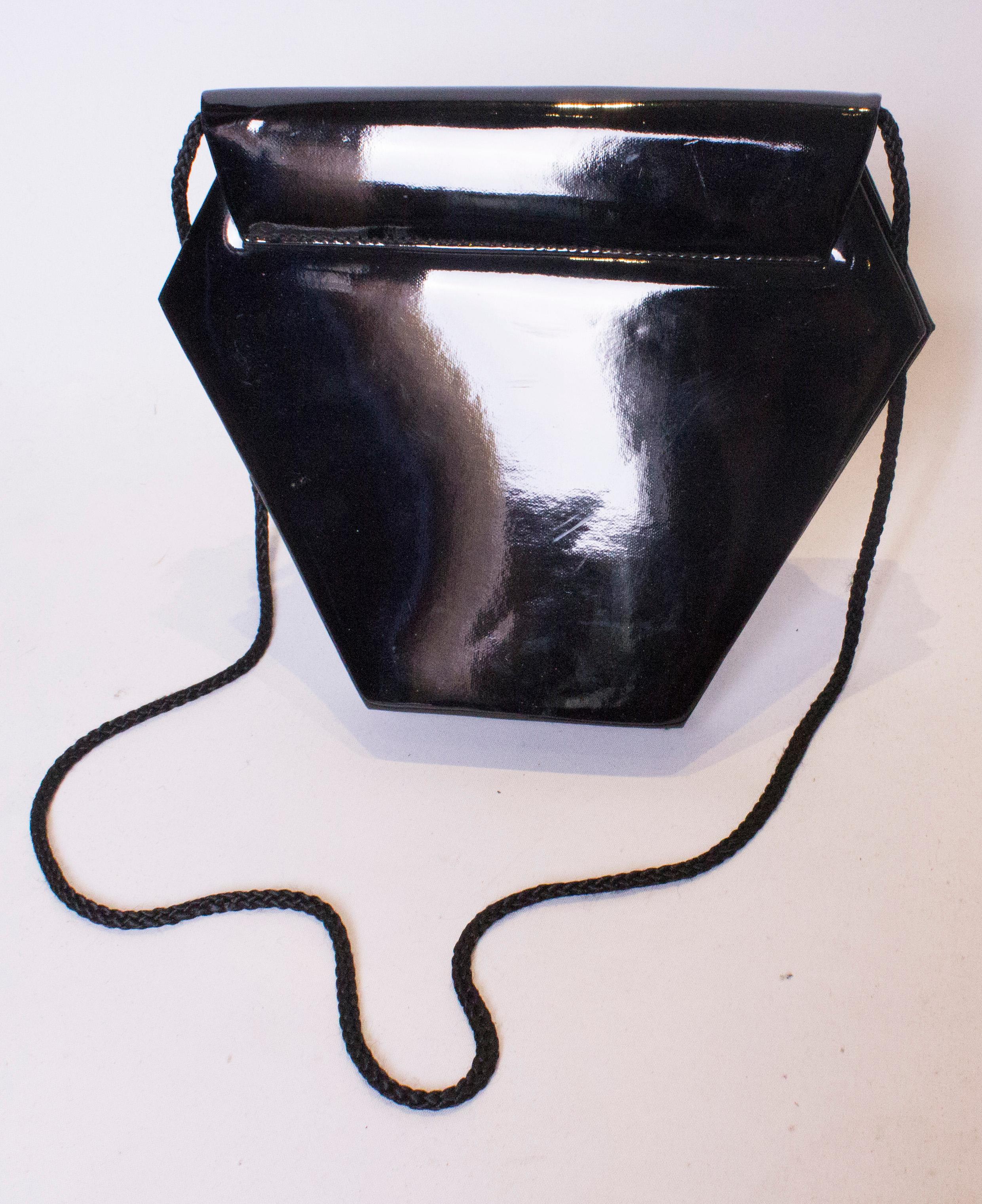 Eine ungewöhnliche schwarze Lacktasche in Hexaganalform. Die Tasche hat eine Klappe auf der Vorderseite mit Druckknopfverschluss, eine Innentasche und einen Schulterriemen mit Seil.