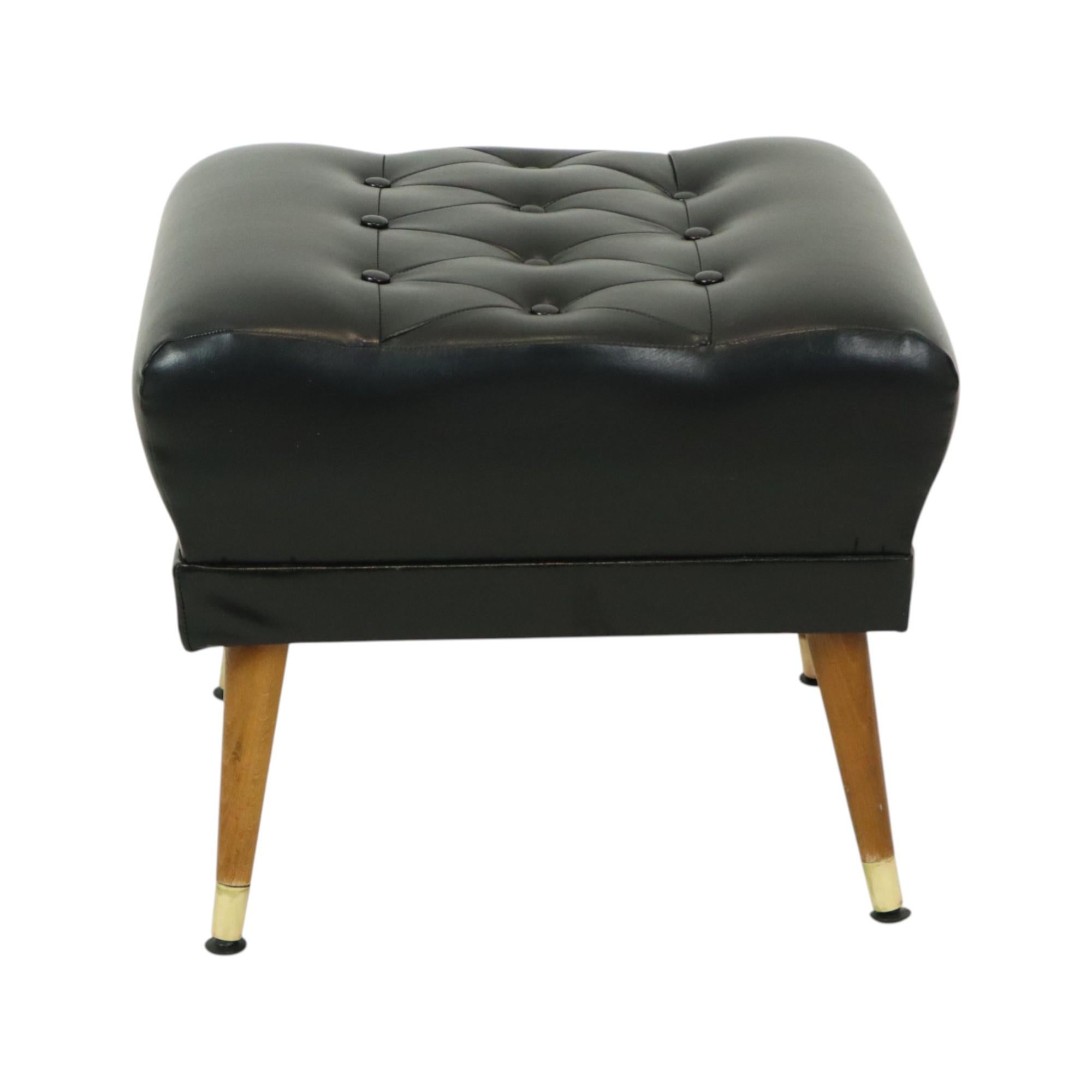 Incantevole pouf vintage rivestito in ecopelle nera di alta qualità (o finta pelle), con seduta imbottita nell'iconico stile Chesterfield. La base è costituita da gambe coniche in legno rifinite con dettagli in ottone. Questo articolo risale alla