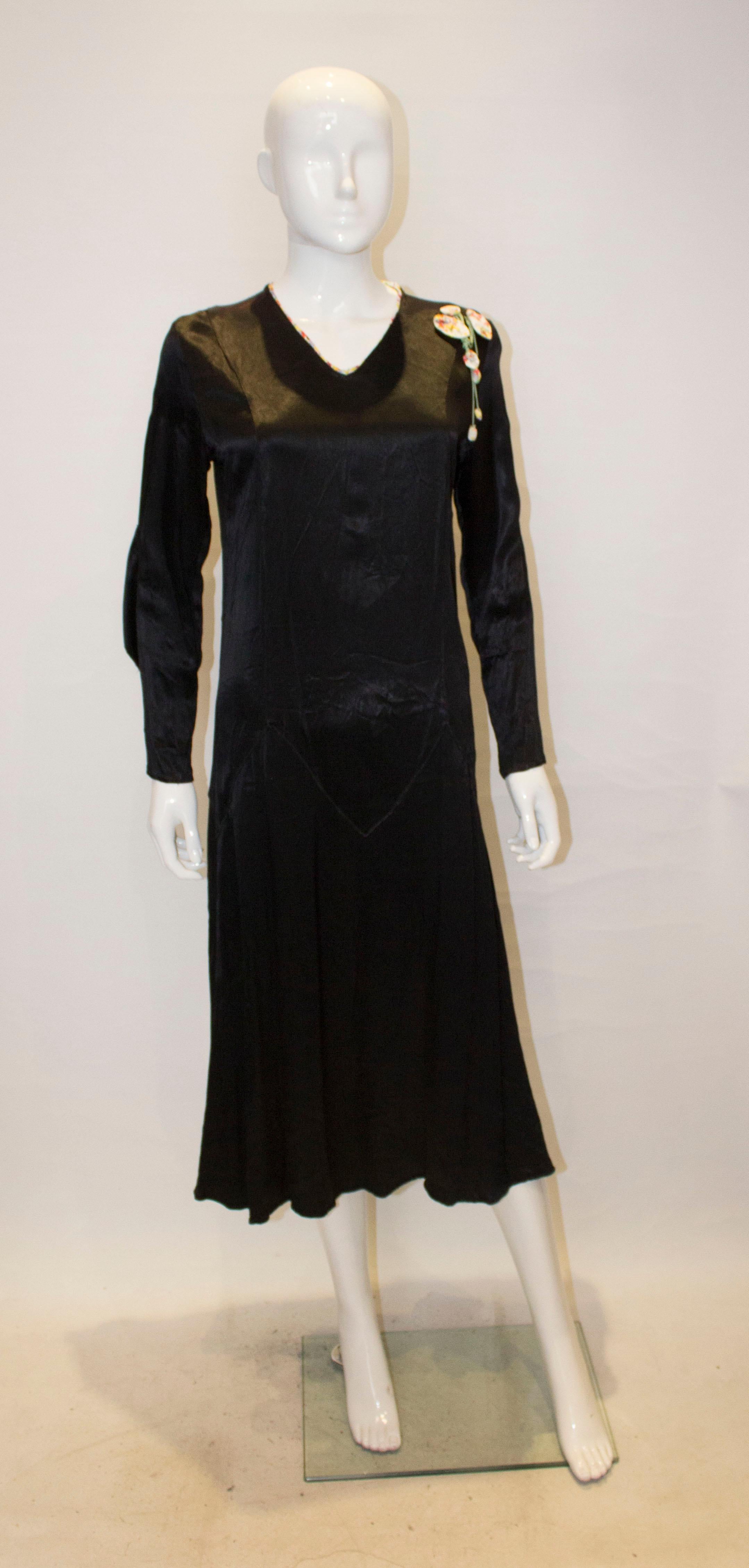 Ein charmantes Vintage-Kleid aus den 1920er Jahren. Das Kleid ist aus schwarzem Satin, hat einen V-Ausschnitt mit Blumenbesatz, Ärmel mit Blumenrüschen und Blumenverzierung an einer Schulter. 