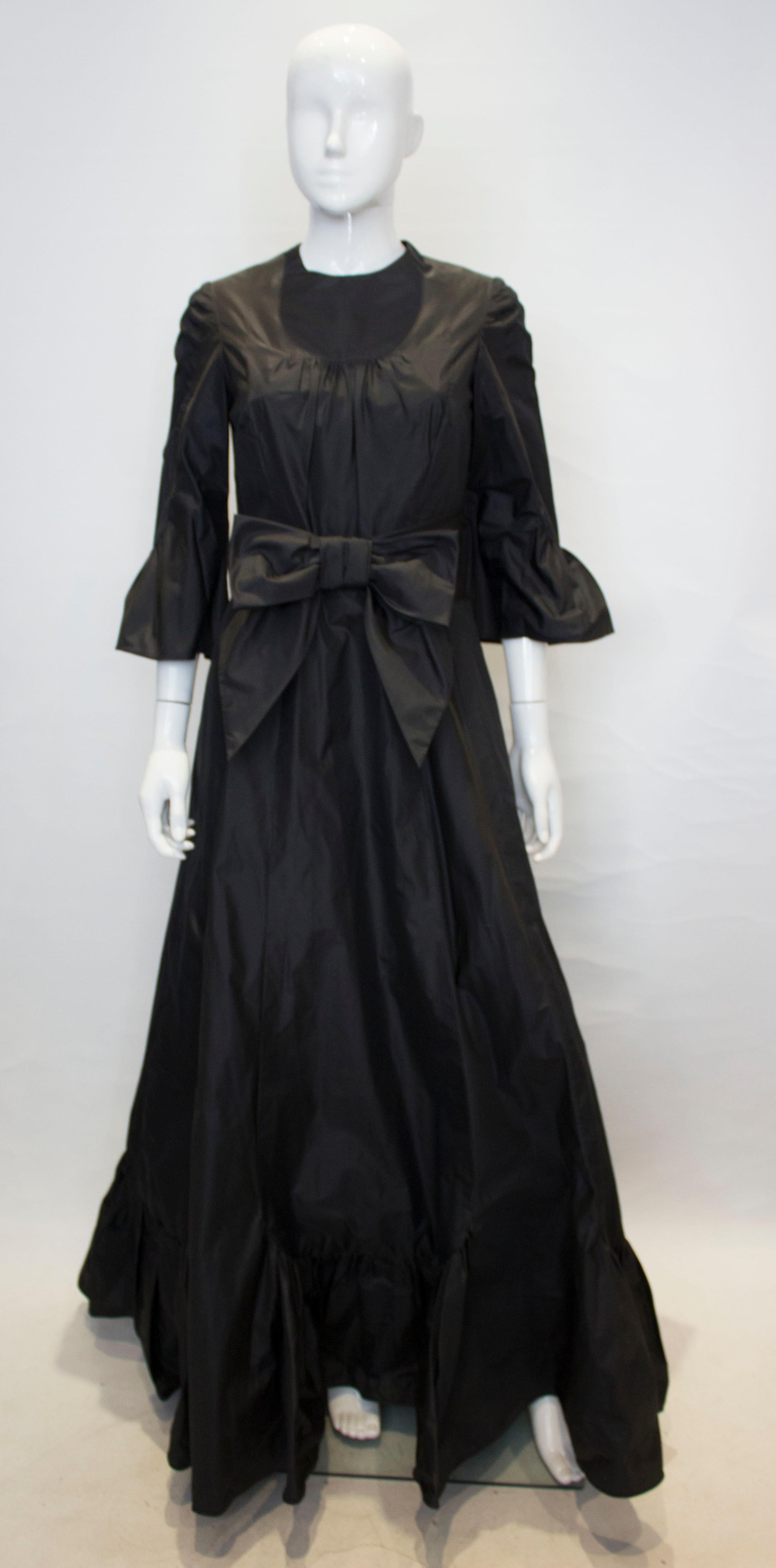 A head turning black silk taffeta gown by Hartnell. The dress has a round neckline, elbow length sleaves with a frill detail and a frill at the hem.  It has a central back zip and bow detail at the waist.
