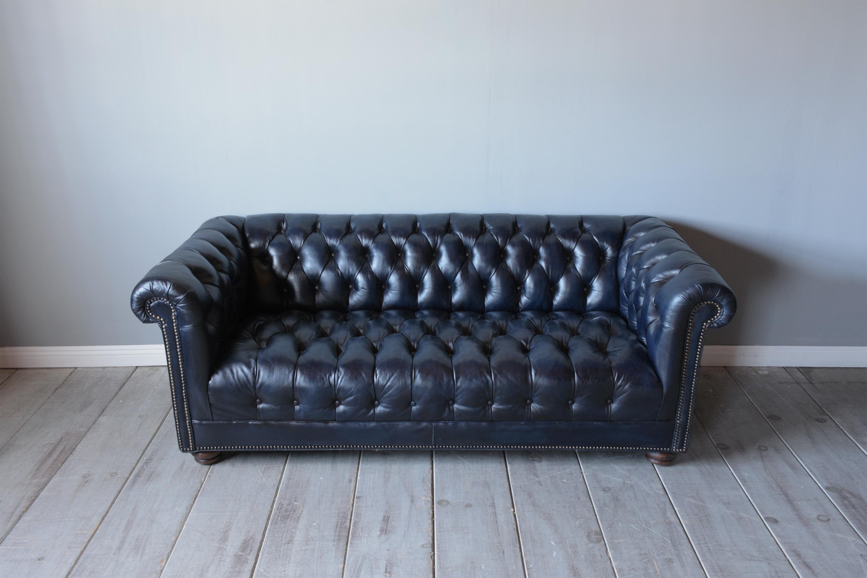 This vintage chesterfield sofa is in great condition, is newly restored, and features its original leather upholstery professionally dyed a dark blue color. This sofa comes with a tufted button design, brass nail details, and rests on walnut bun