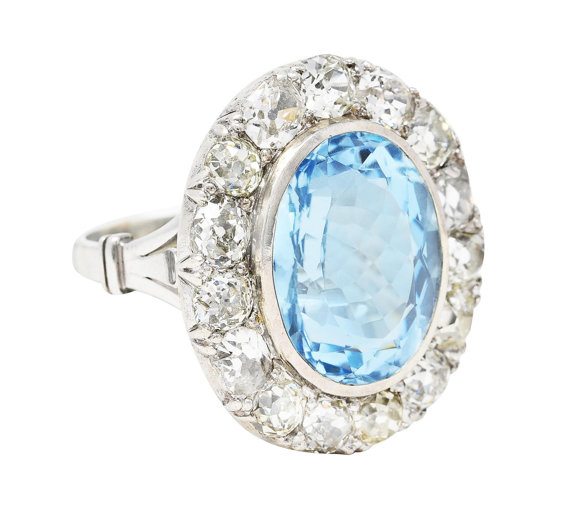 Cluster ring features a mixed oval cut blue topaz measuring 15.5 x 12.2 mm. Transparent with light greenish blue color. Bezel set in polished white gold and surrounded by old mine cut diamonds. Total diamond weight is approximately 3.75 carats. Near
